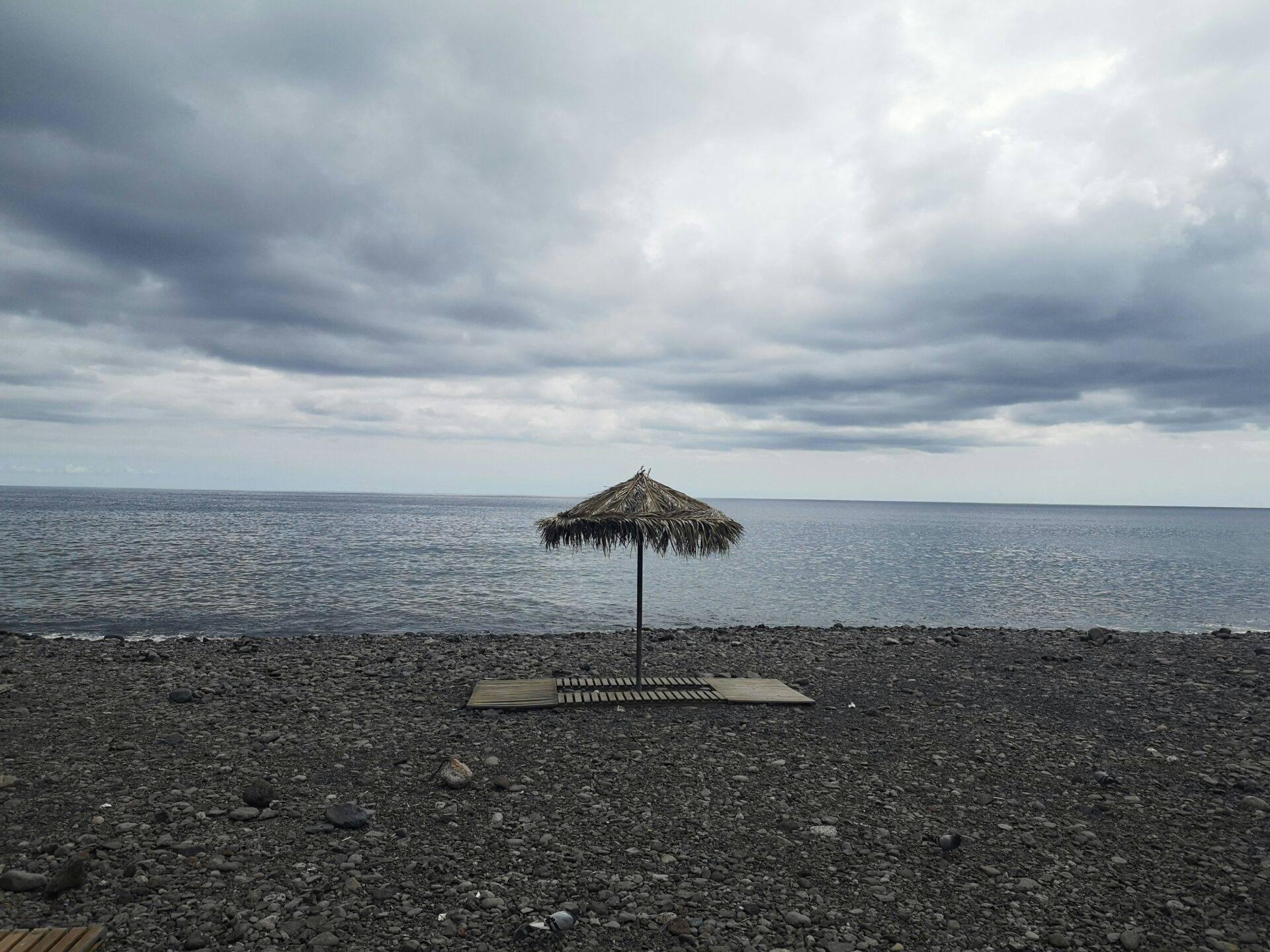 Un parasol de paille solitaire sur une plage de galets gris face à une mer calme sous un ciel nuageux