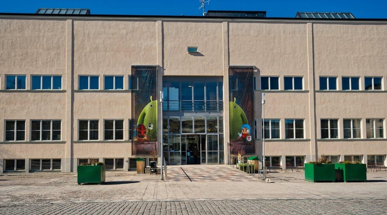 Moderne Fassade eines institutionellen Gebäudes mit verglastem Eingangsbereich und farbenfrohen Kindermotiven auf den seitlichen Tafeln