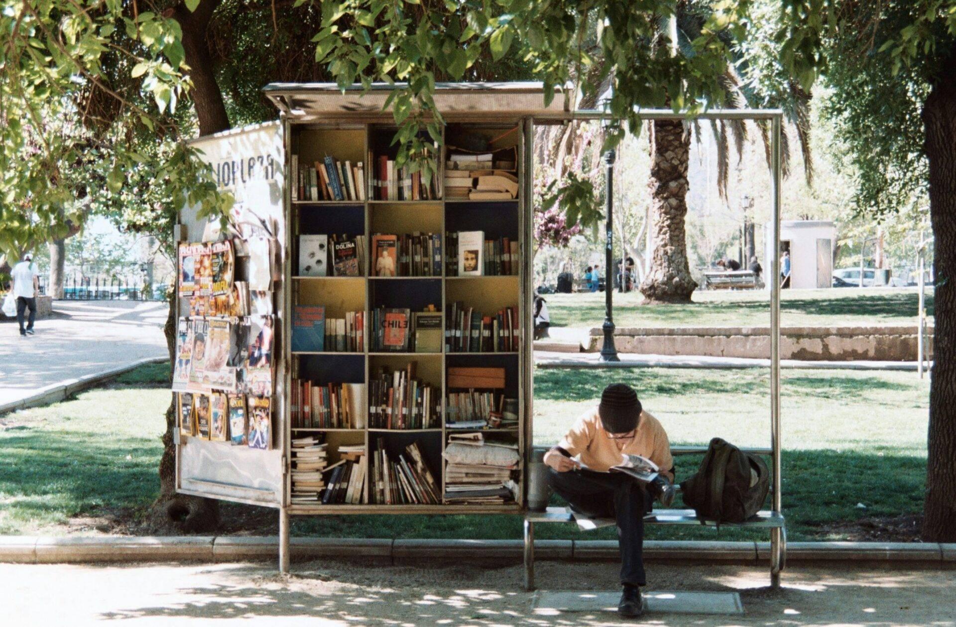 Ein Mann liest sitzend vor einer kleinen Freiluftbibliothek unter Bäumen in einem Stadtpark