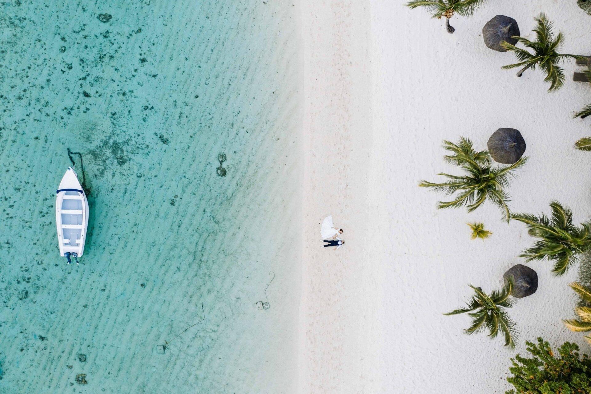 Vista aérea de una playa tropical con un barco blanco fondeado, arena blanca y palmeras