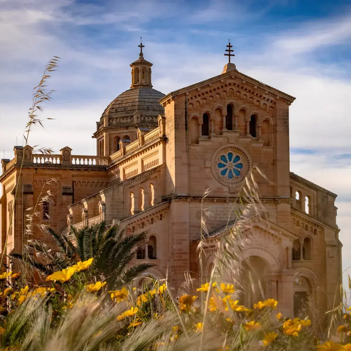Beigefarbene Steinkathedrale mit Kuppel und Kreuz, umgeben von gelben Blumen und Palmen unter blauem Himmel