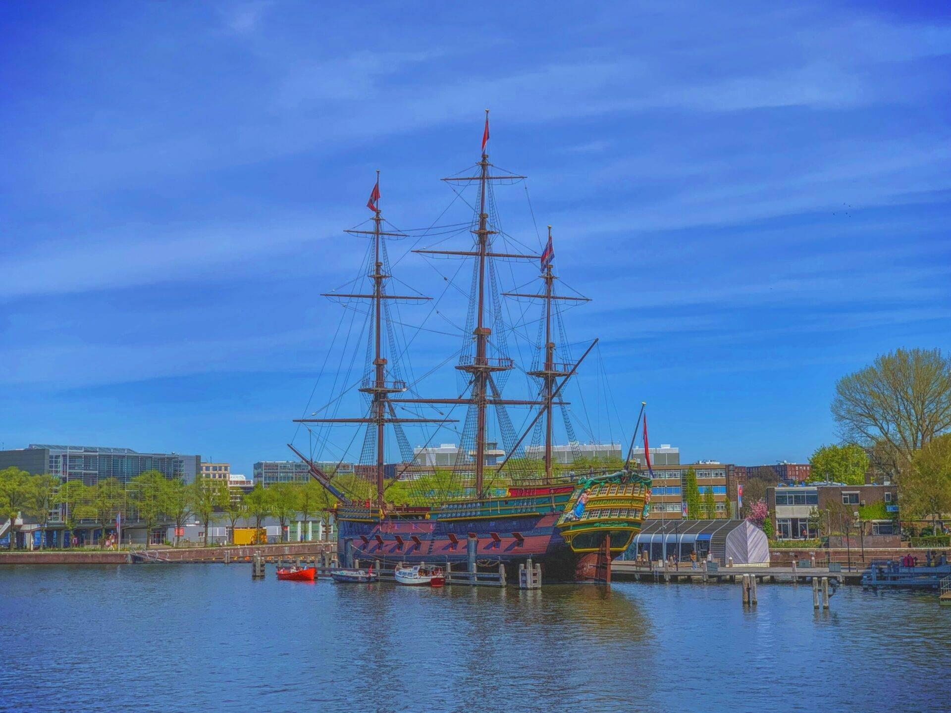 Ein buntes Dreimaster-Segelschiff liegt in einem städtischen Flusshafen unter klarem blauen Himmel