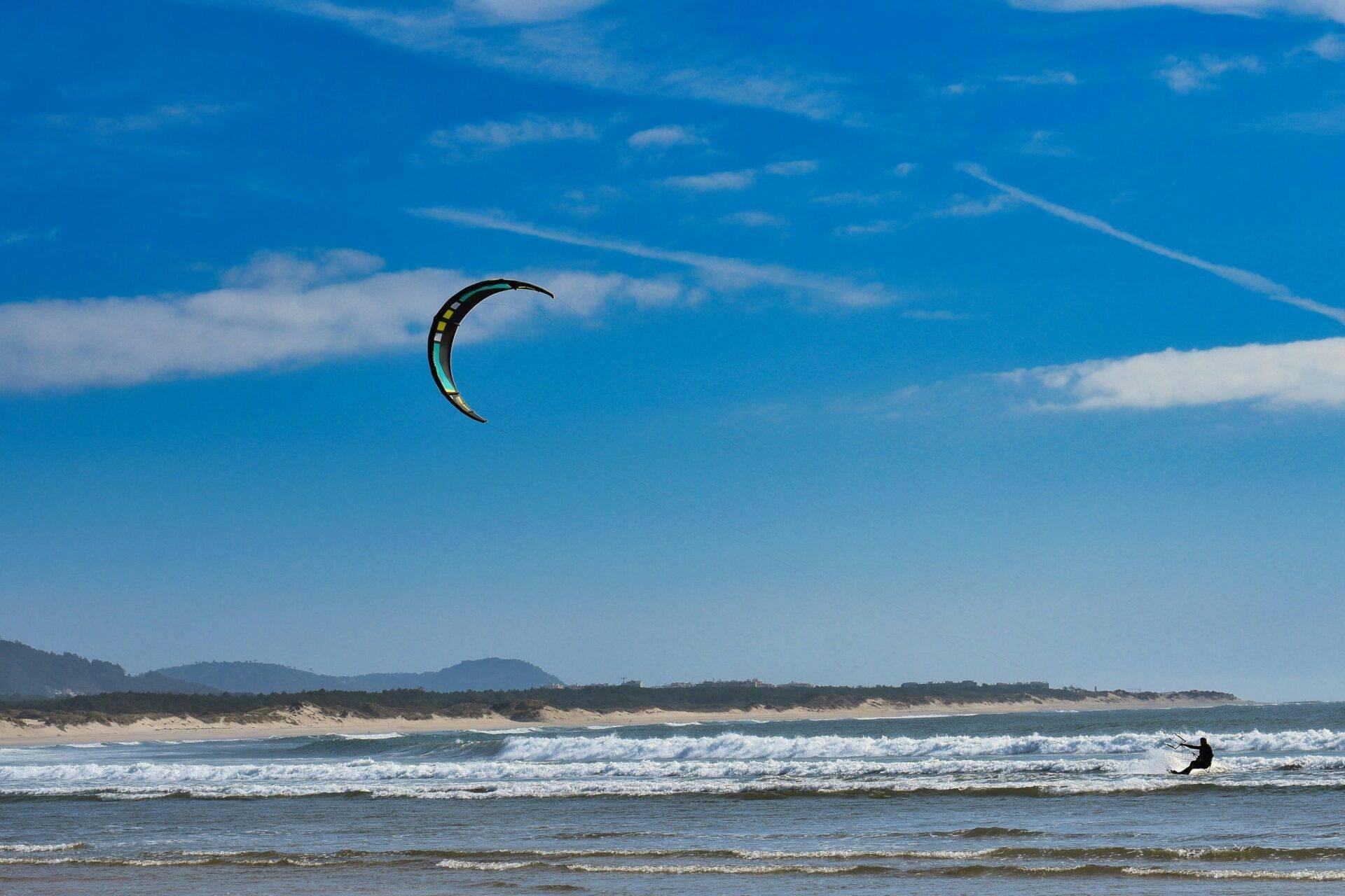 Ein Kitesurfer in Aktion auf einem feinen Sandstrand mit Wellen, unter blauem Himmel und Bergen im Hintergrund