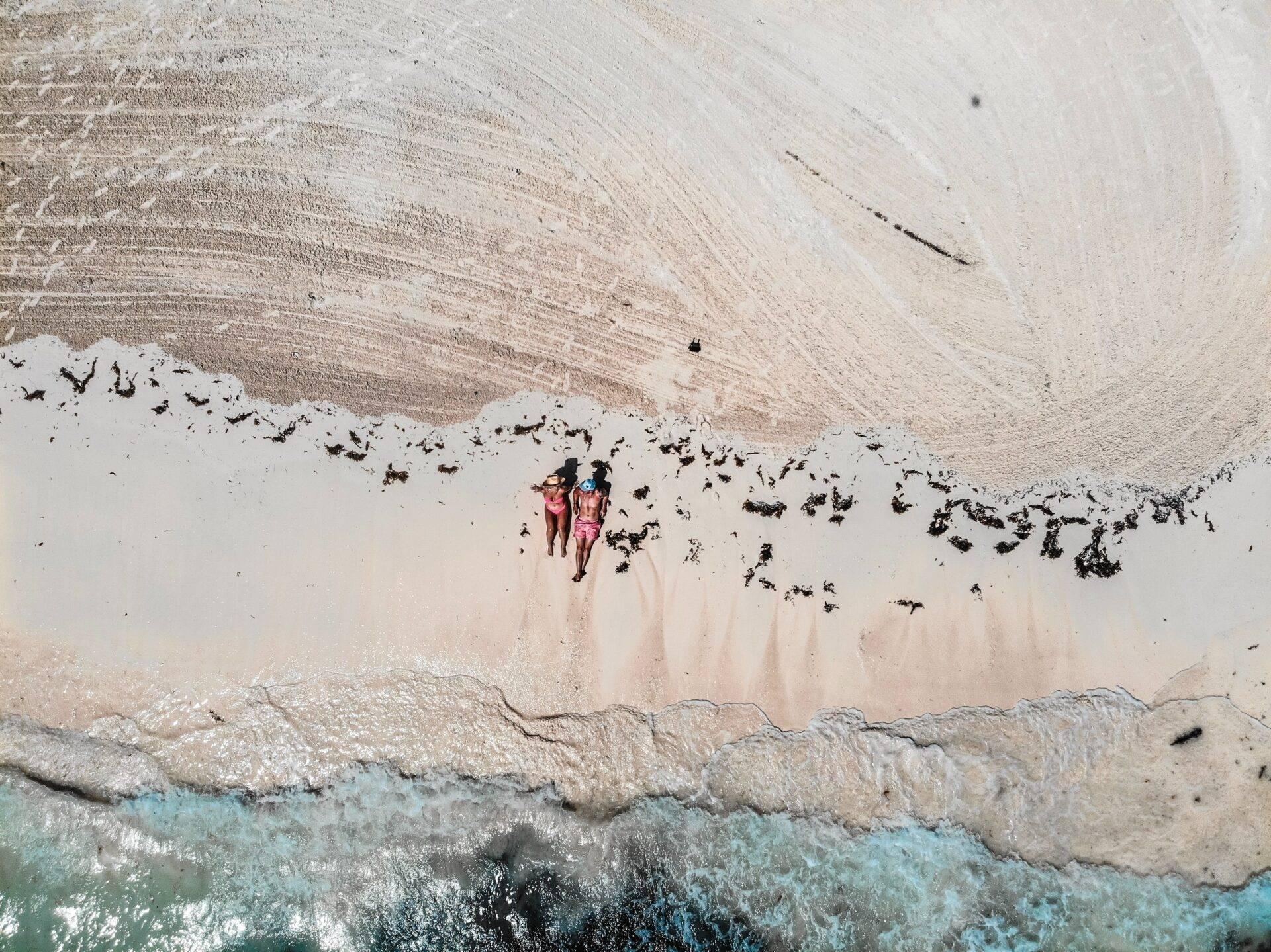Two people in swimwear on a white sand beach facing crystal-clear turquoise waves