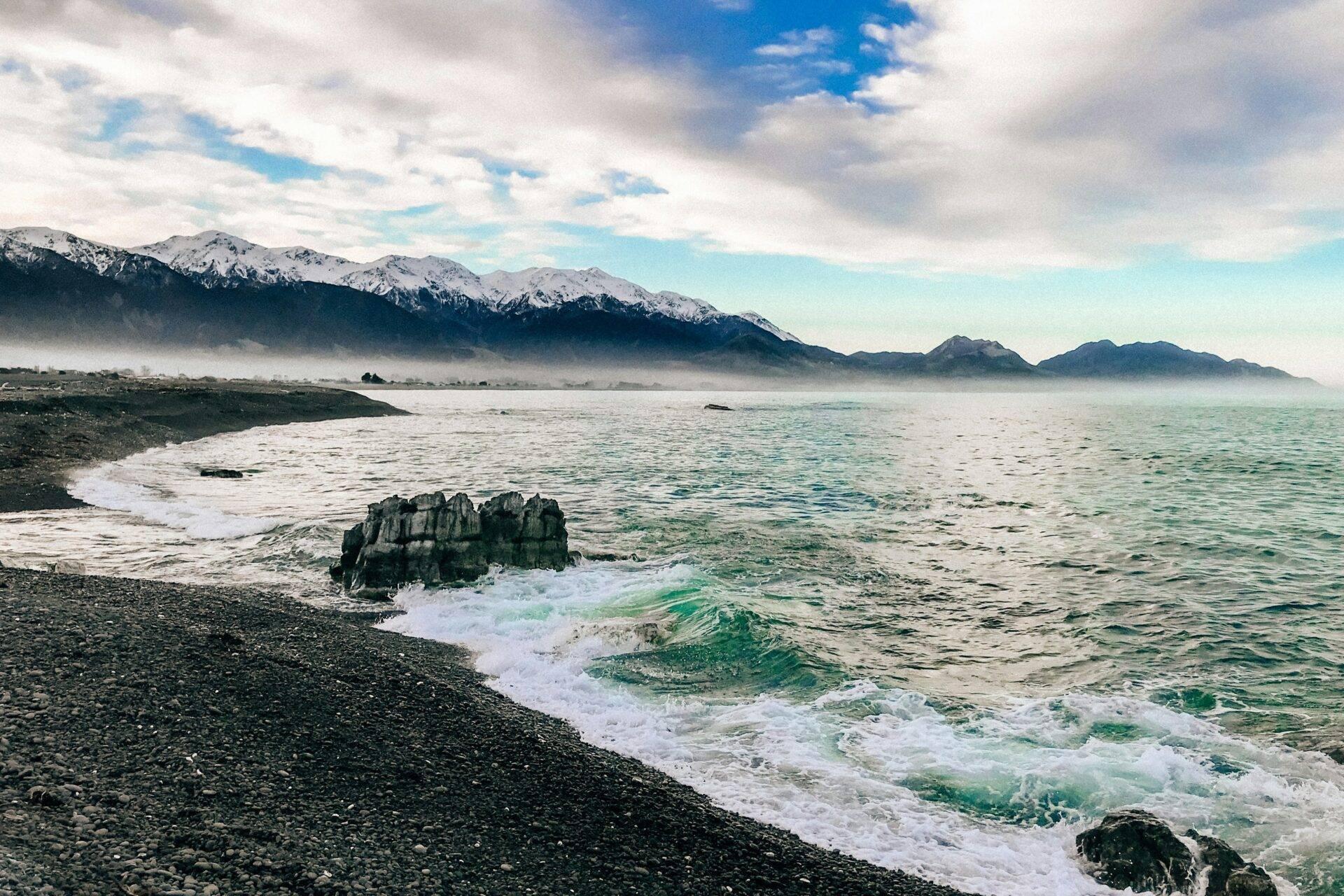 Costa rocciosa con onde turchesi, montagne innevate sullo sfondo e nebbia mattutina sulla baia