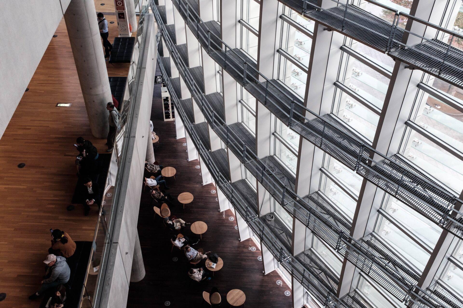 Luftaufnahme eines modernen Atriums mit Glasfront, in dem Besucher an runden Tischen entlang einer Promenade sitzen