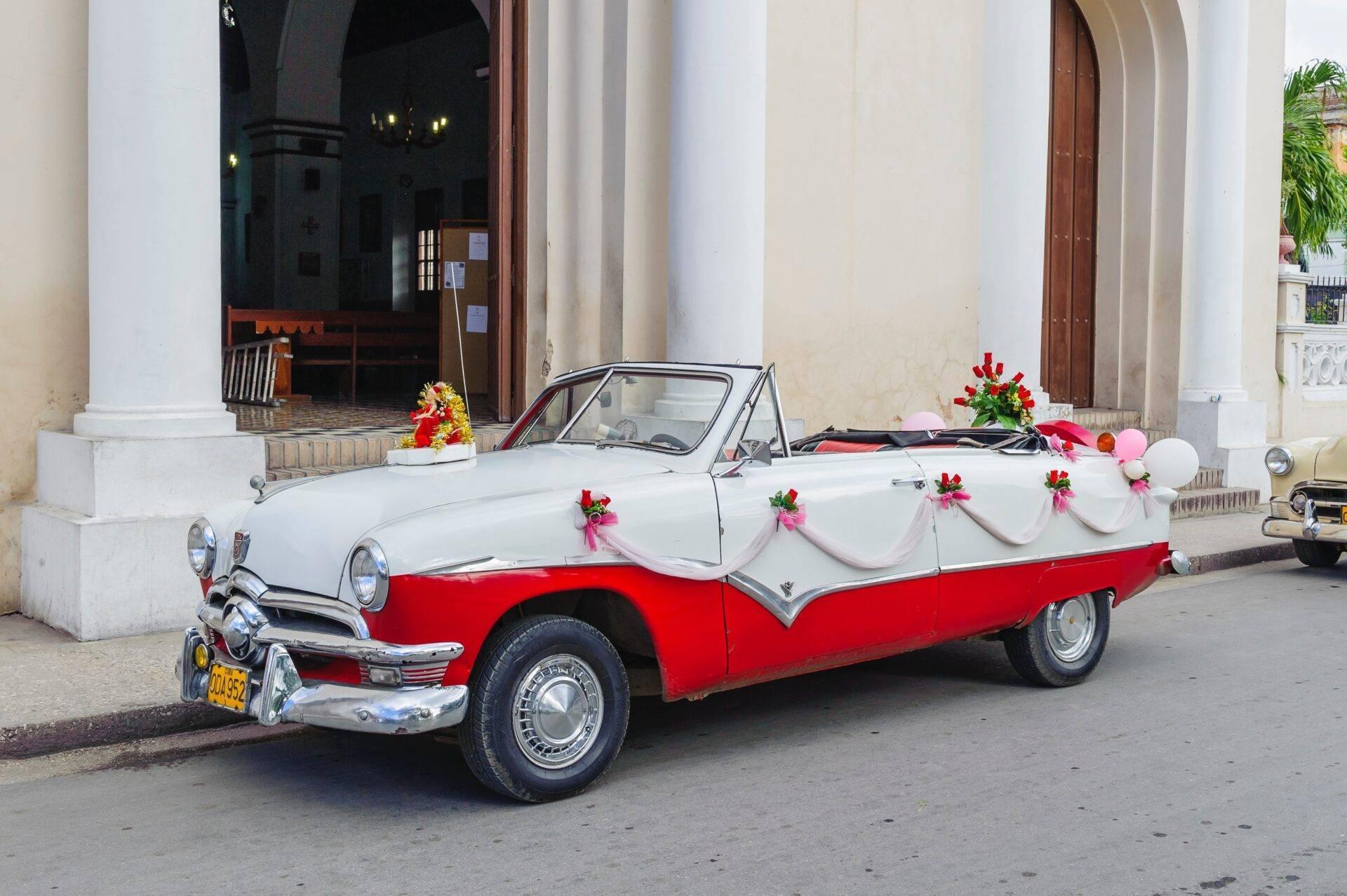 Un’auto decapottabile rossa e bianca decorata con fiori rosa parcheggiata davanti a un edificio coloniale bianco