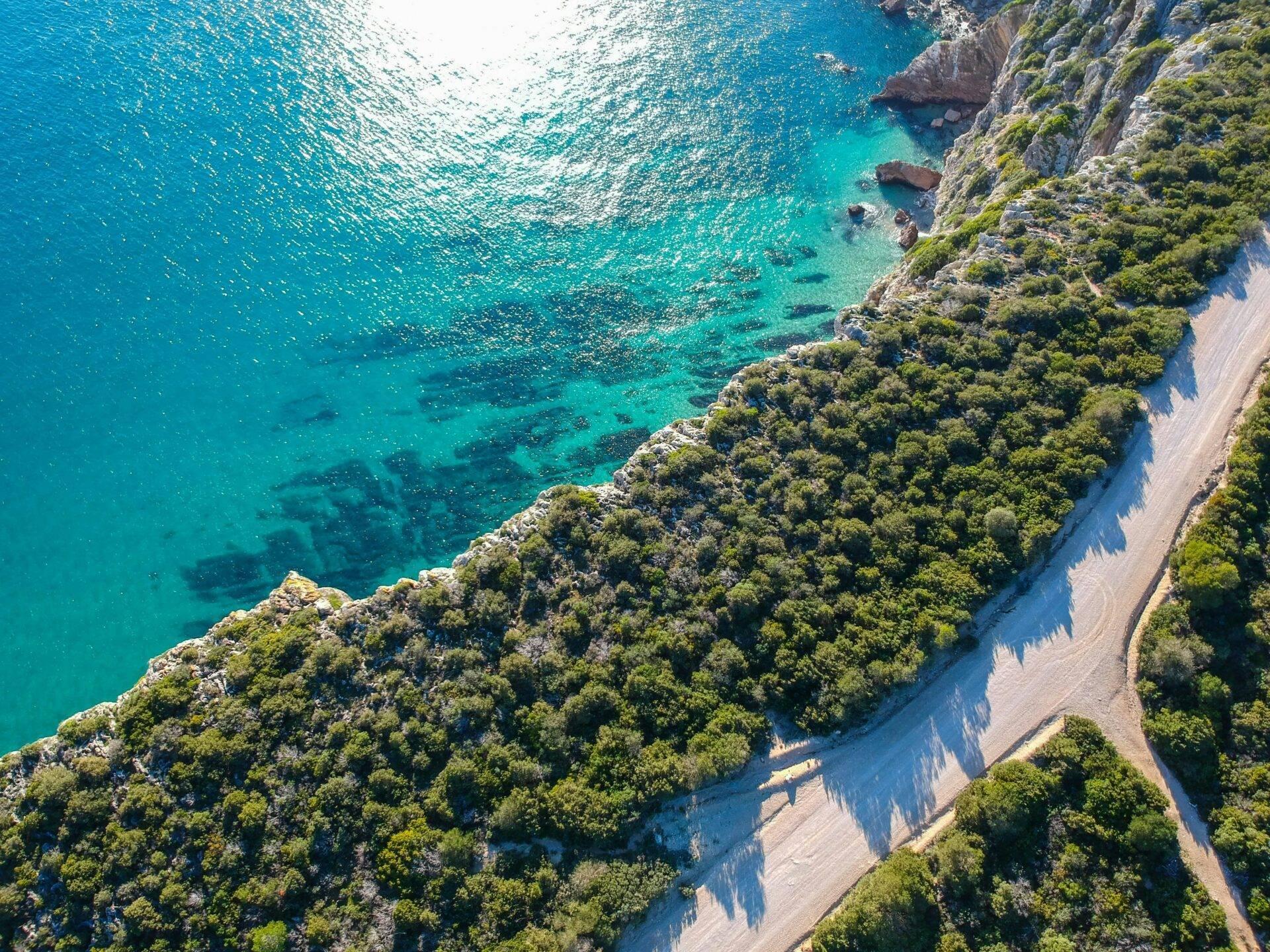 Luftaufnahme einer Mittelmeerküste mit bewaldeten Klippen, Sandstrand und kristallklarem türkisfarbenem Wasser