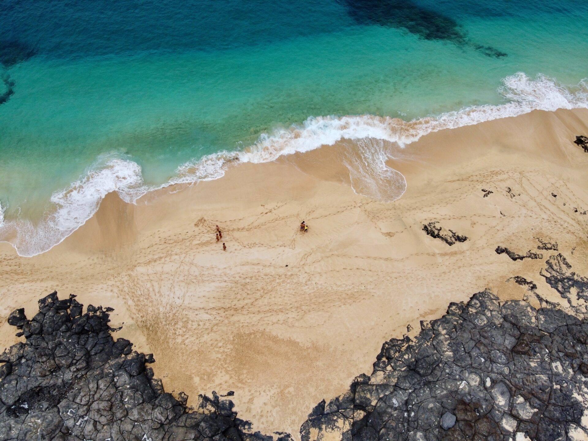 Vista aerea di una spiaggia di sabbia fine con onde turchesi e rocce nere che incorniciano una riva deserta