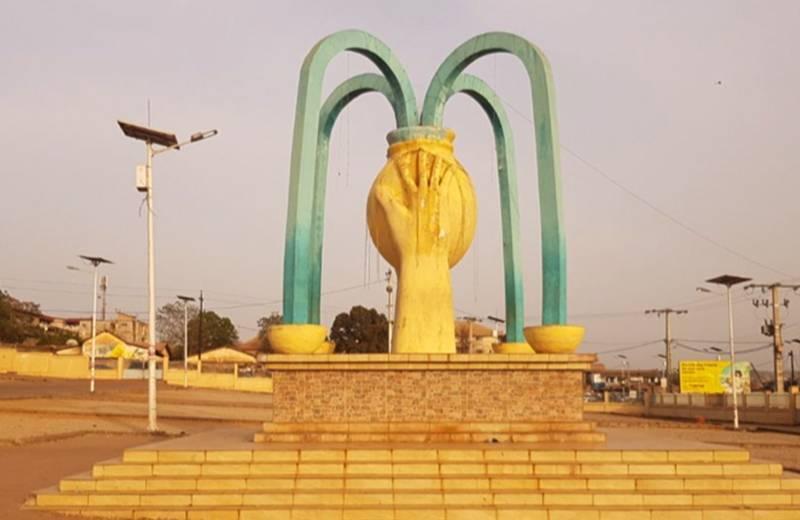Monumento pubblico con archi verdi e mano gialla centrale in una piazza urbana assolata
