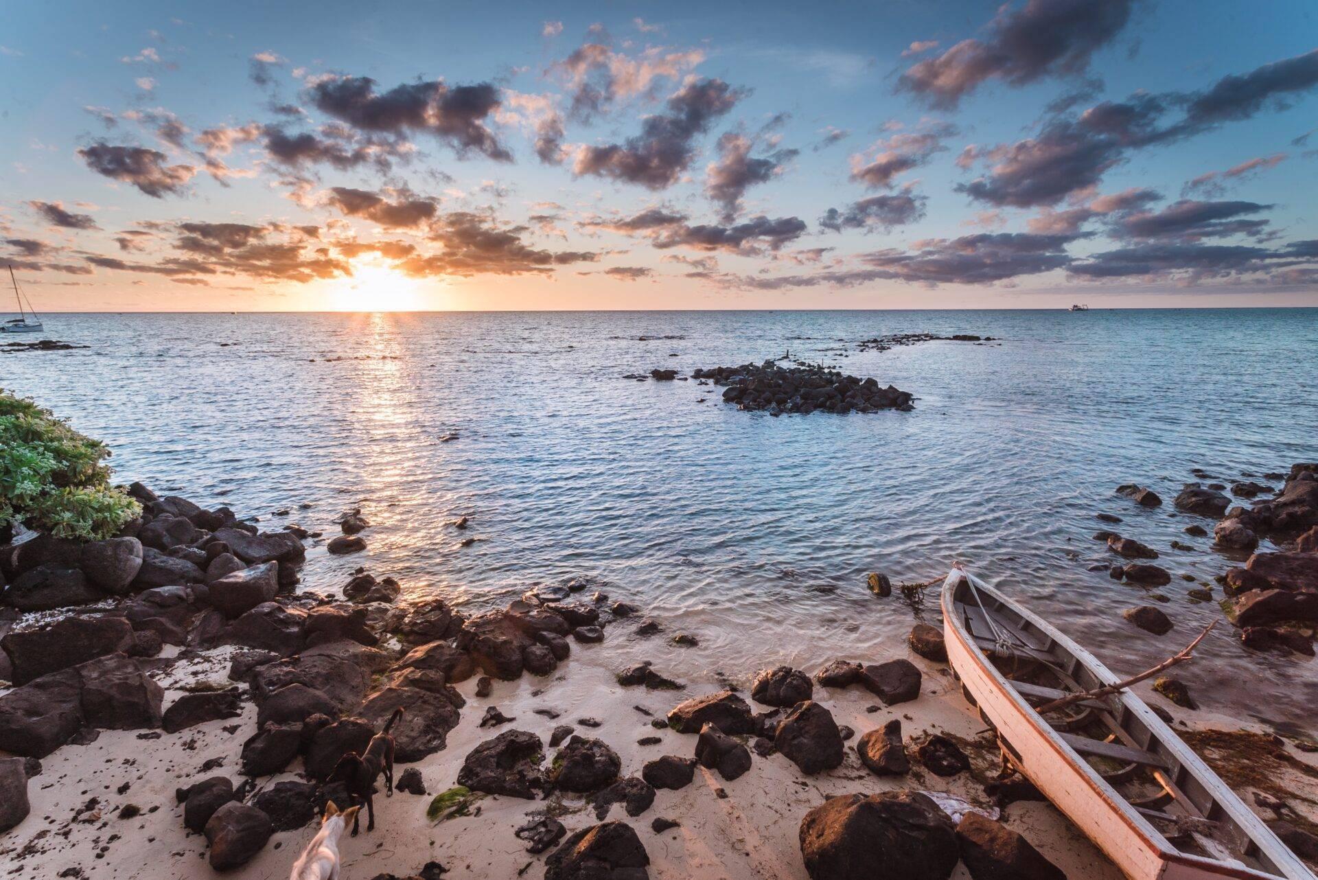 Atardecer sobre una playa rocosa con dos barcas de pesca varadas y veleros al fondo