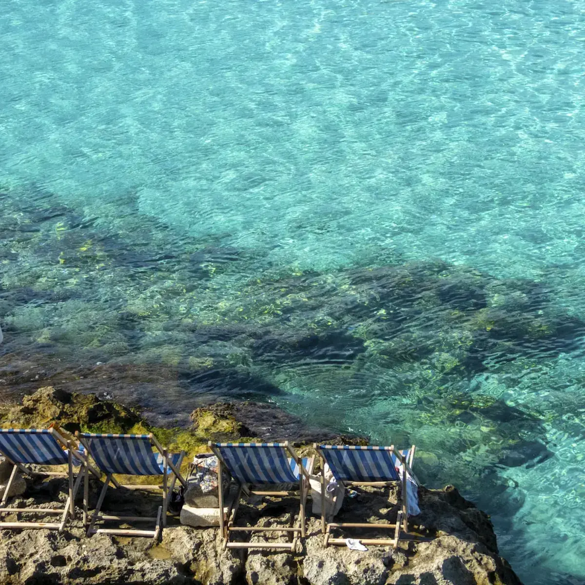 Blau-weiß gestreifte Liegestühle auf Felsen vor kristallklarem mediterranem Meer