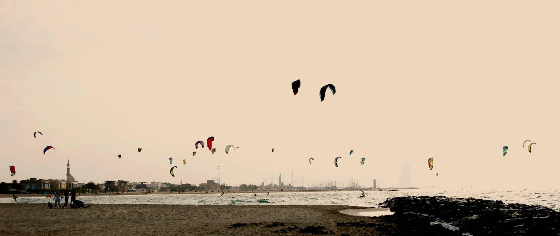 Ein Strand mit zahlreichen Kitesurfern im Wasser und vertäuten Segelbooten vor einer Küstenstadt
