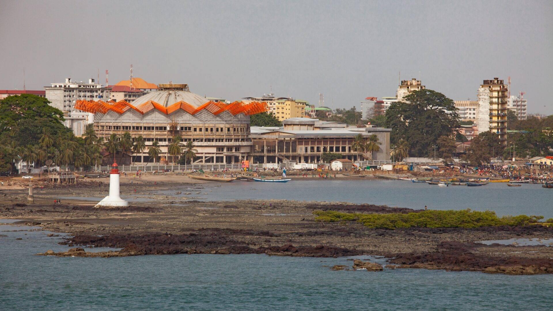 Vista costiera di una città africana con un edificio dai tetti ondulati arancioni, un faro bianco e barche da pesca