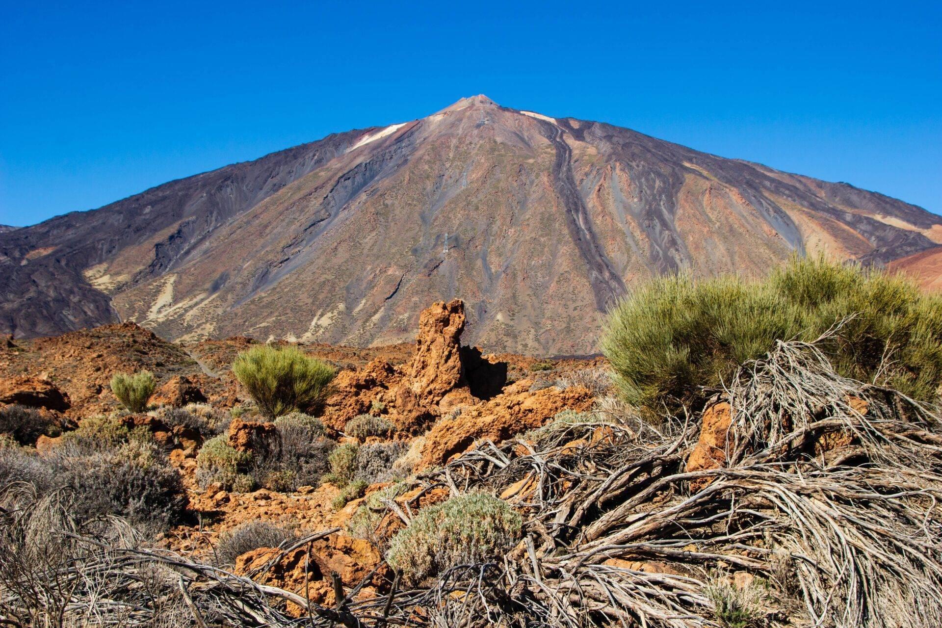 Un vulcano arido con versanti striati di minerali colorati, circondato da una rada vegetazione desertica