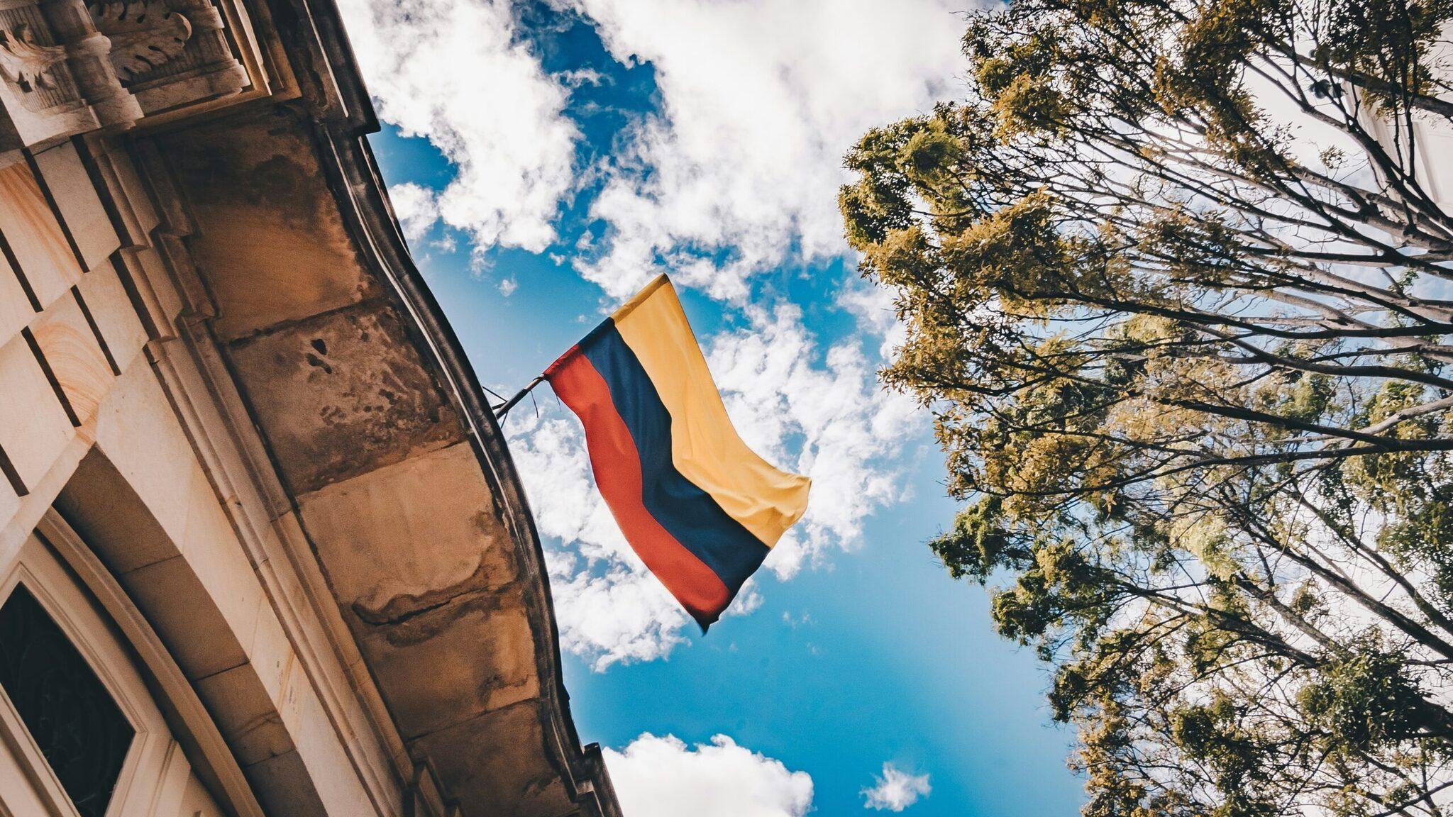 Bandera colombiana ondeando al viento frente a una fachada colonial bajo un cielo azul despejado