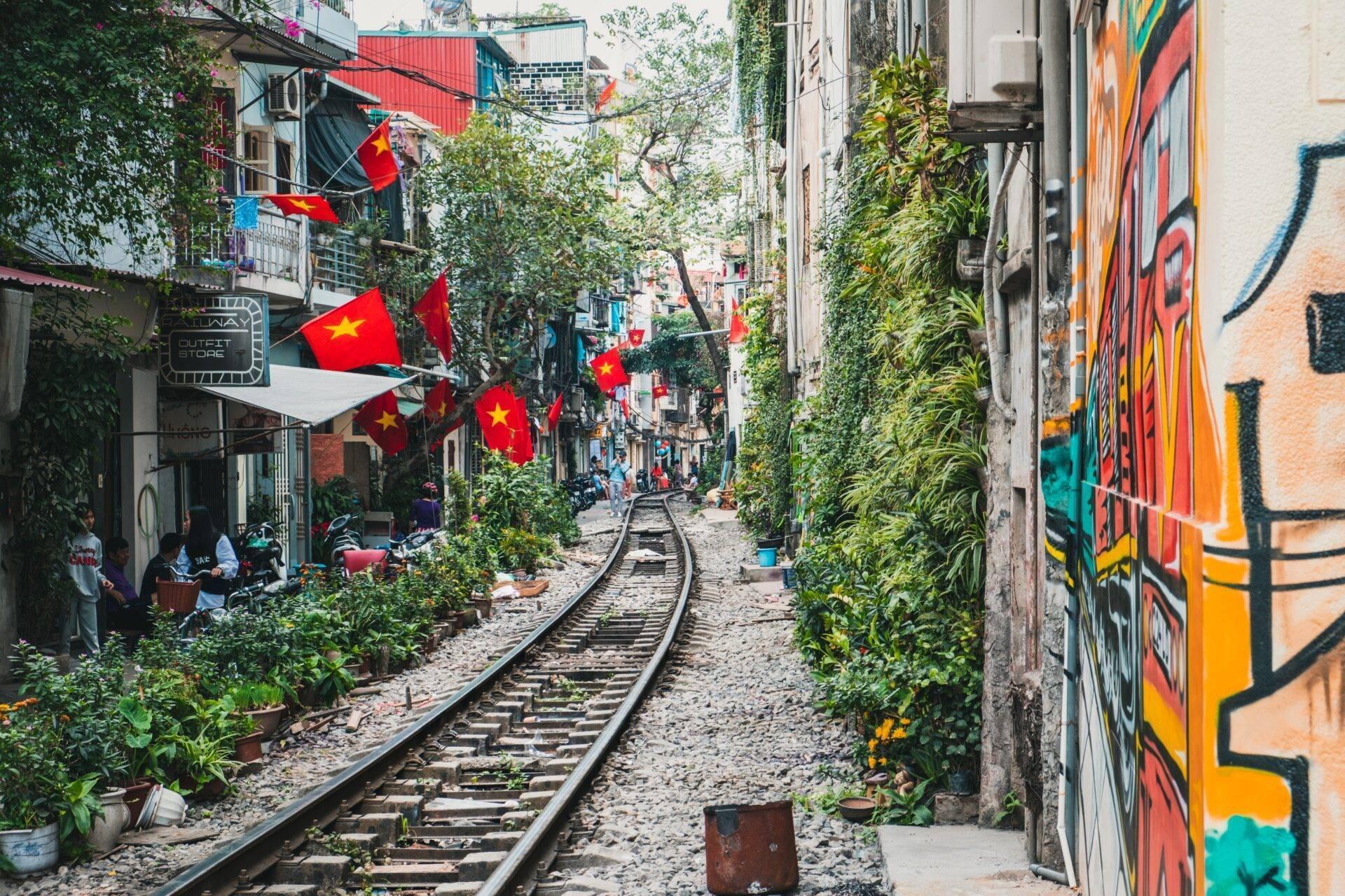 Un callejón estrecho con vías de tren, edificios coloridos, vegetación exuberante y banderas vietnamitas rojas