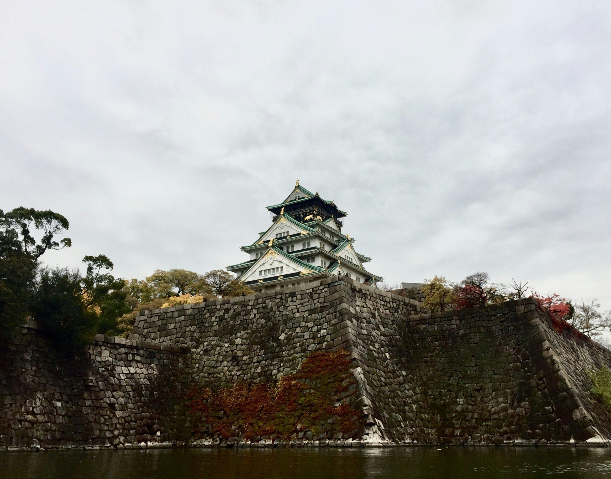 Un castello giapponese dai tetti verdi arroccato su imponenti mura di pietra, che domina l’acqua con vegetazione autunnale