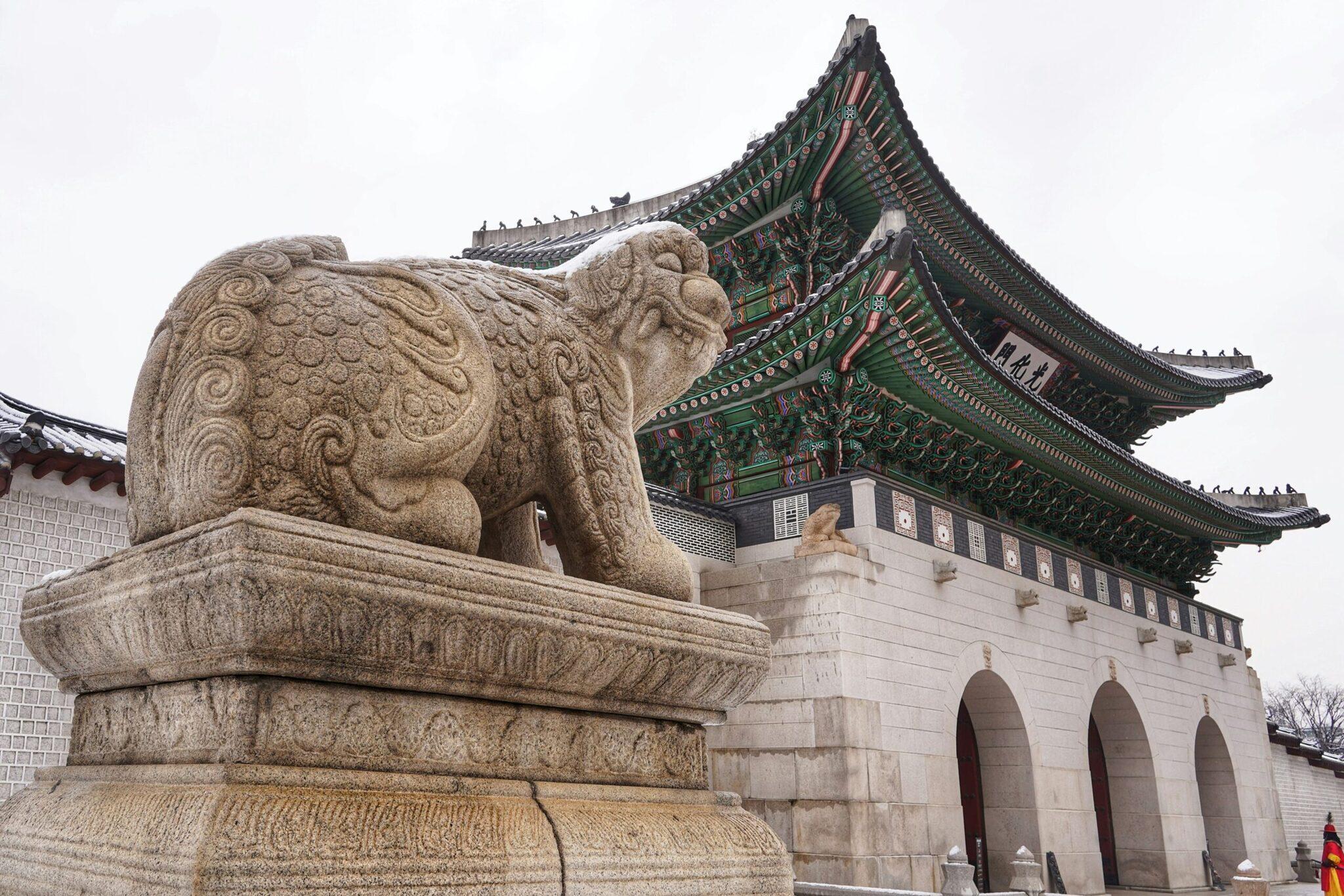 Eine steinerne Löwenskulptur vor einem traditionellen koreanischen Pavillon mit reich verzierten grünen und roten Dächern
