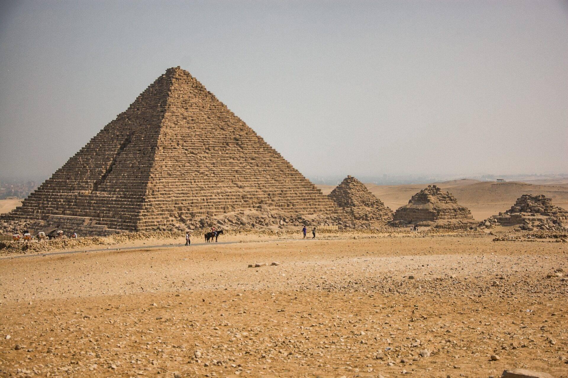 Una grande piramide in pietra nel deserto egiziano con visitatori e rovine circostanti sotto un cielo limpido