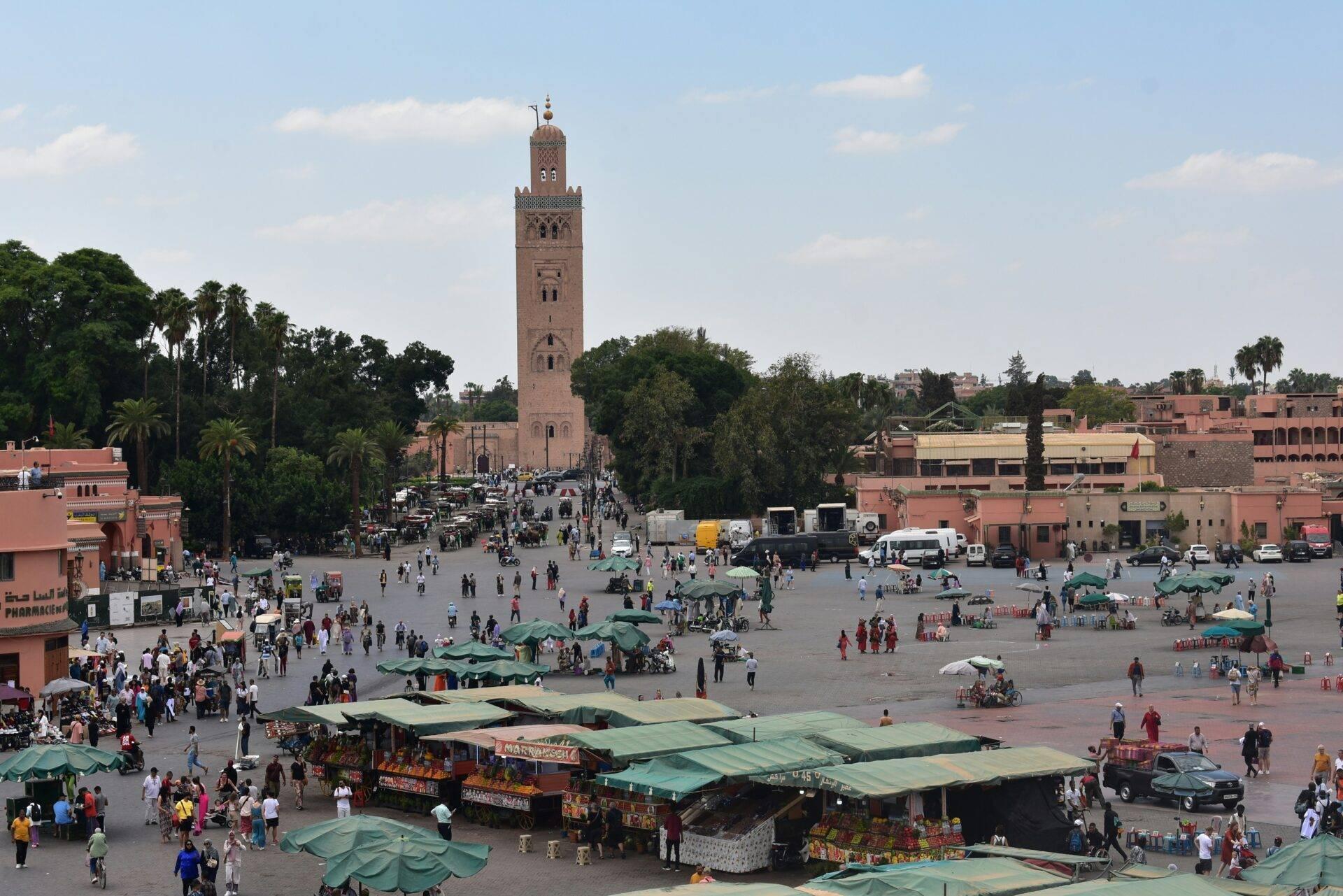Una gran plaza animada con mezquita histórica, vendedores bajo toldos verdes, turistas y edificios de color ocre tradicional