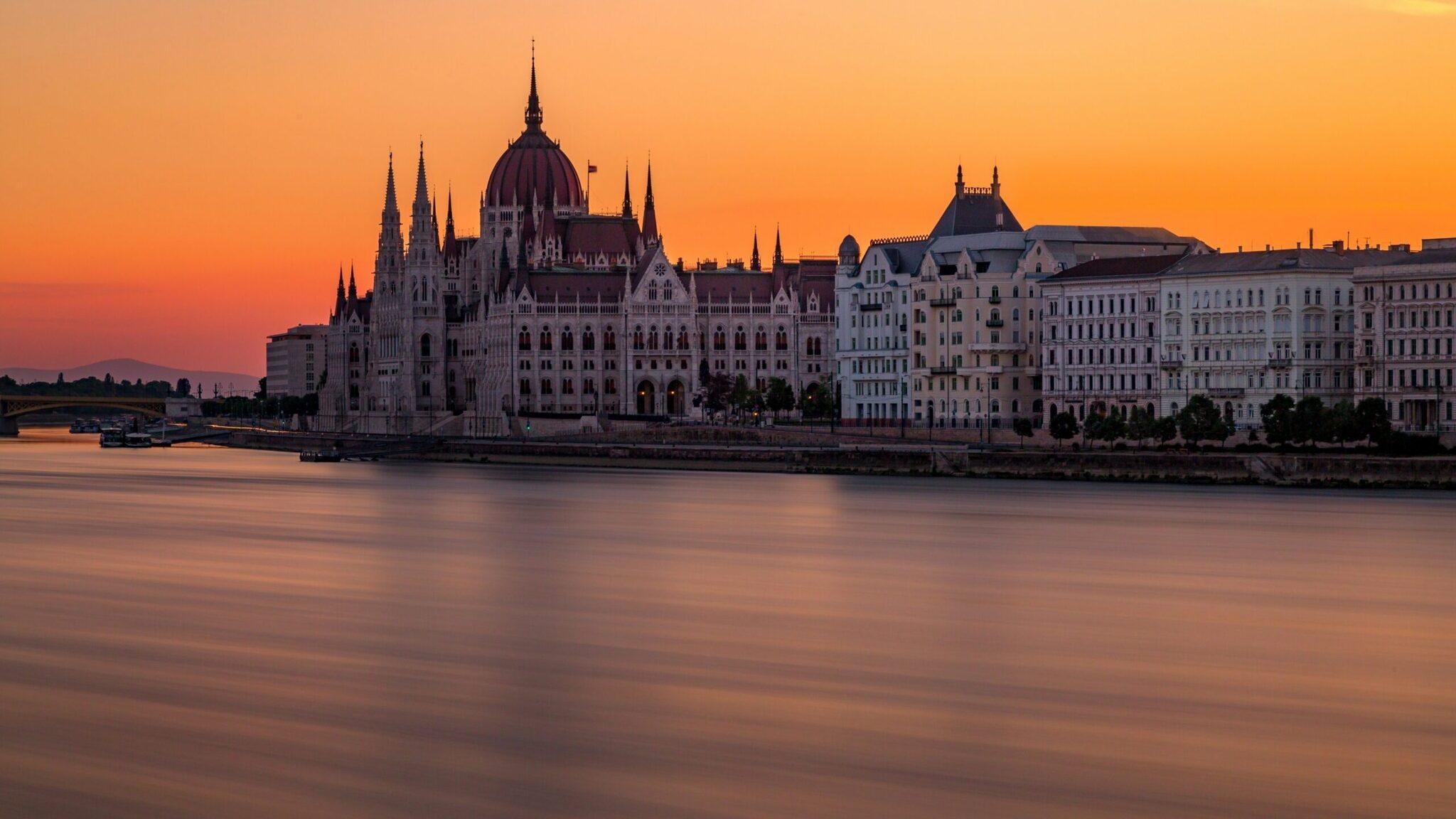 The Budapest Parliament Building floodlit at sunset on the banks of the Danube, its Gothic spires and central dome reflected in the water