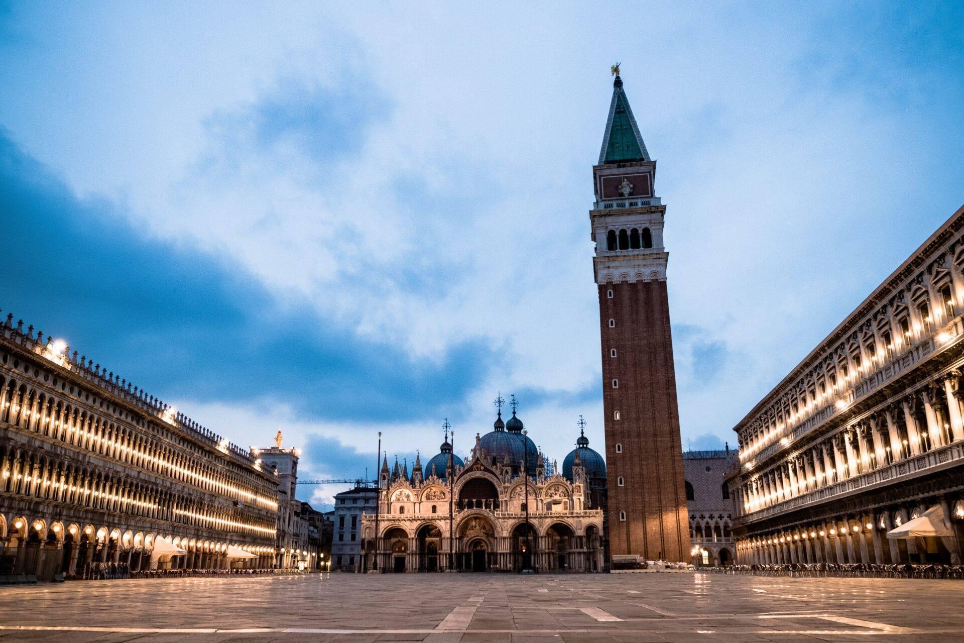 Vasta piazza lastricata circondata da palazzi illuminati con una basilica ornata e un campanile in mattoni rossi