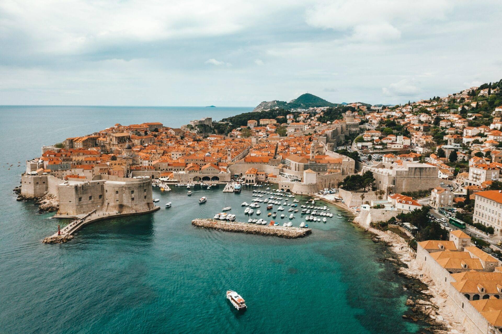 Veduta aerea di una città costiera fortificata con tetti arancioni, porto turistico e montagna sullo sfondo