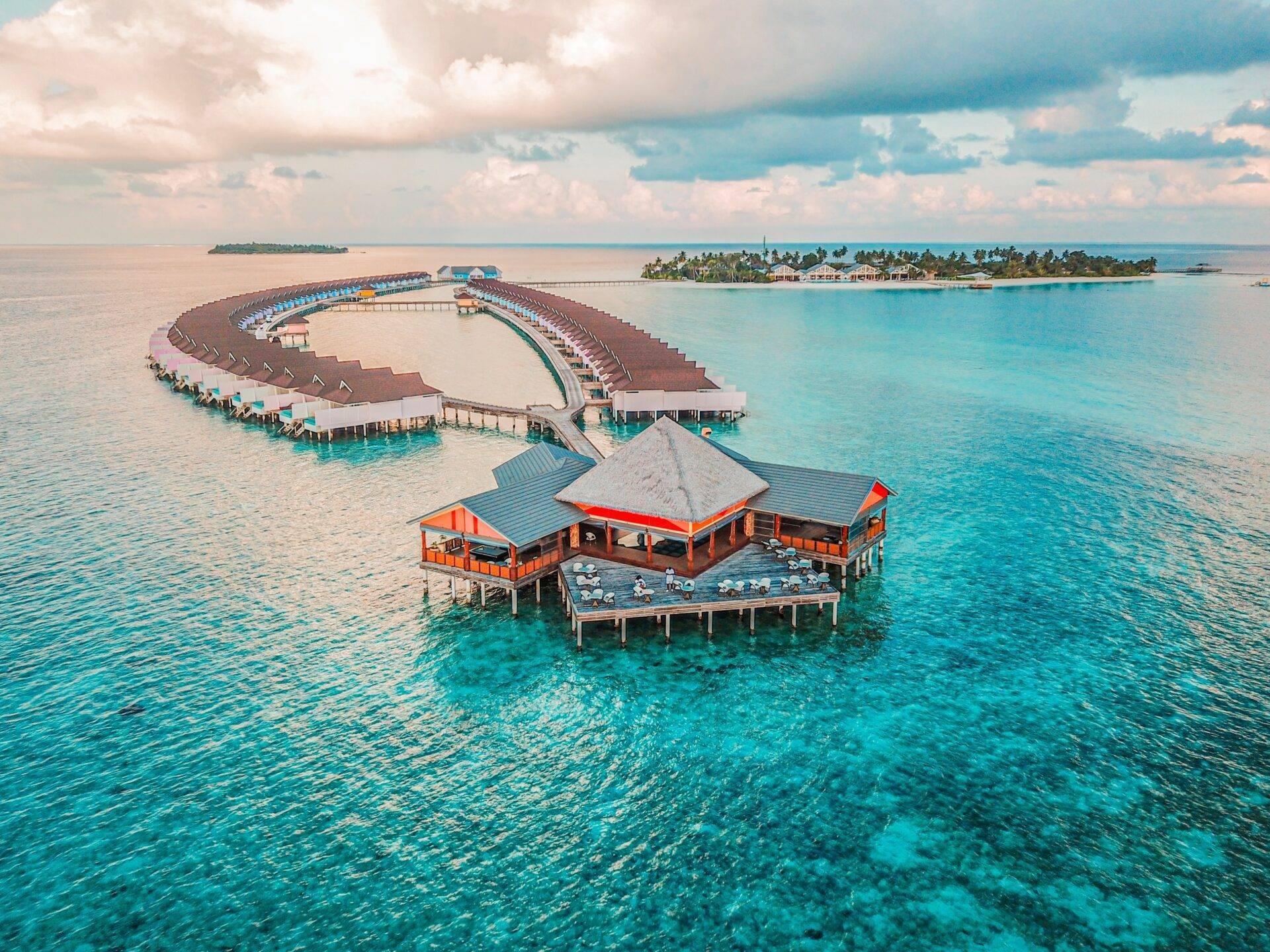 Luftaufnahme eines Tropenresorts mit Überwasserbungalows in Spiralform, türkisfarbenem Lagon und dicht bewaldeten Inseln