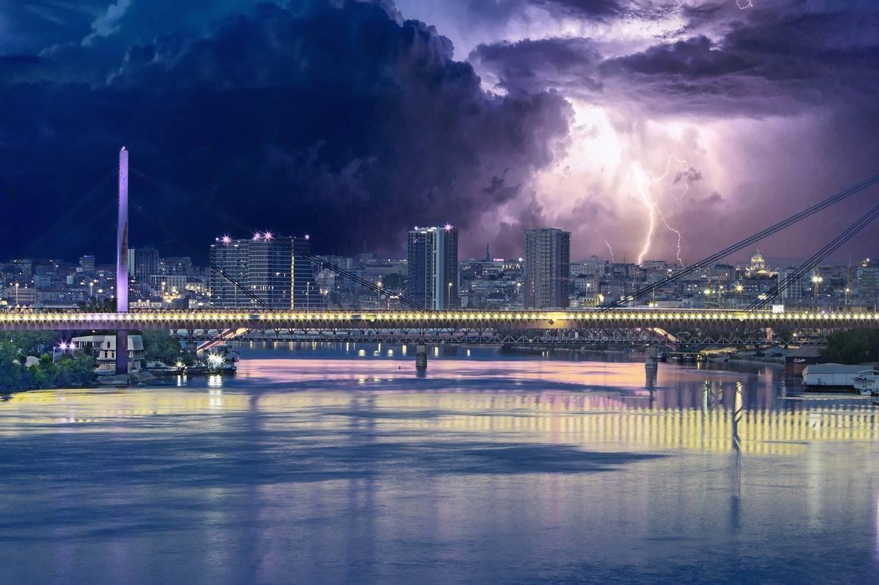 Eine beleuchtete Brücke über einen Fluss mit einer Stadt im Hintergrund unter einem spektakulären Gewitter