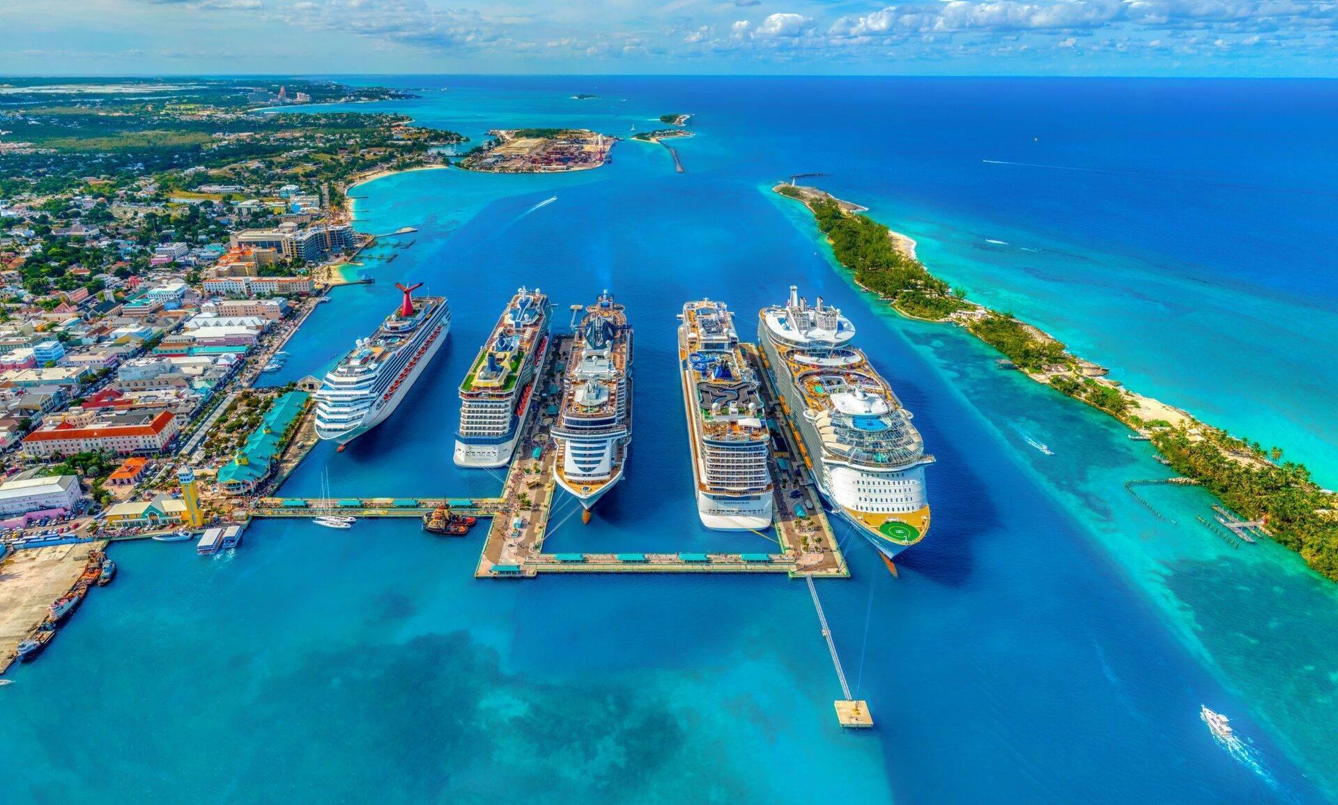 Aerial view of a tropical cruise port with six ships docked in a turquoise bay surrounded by green islands