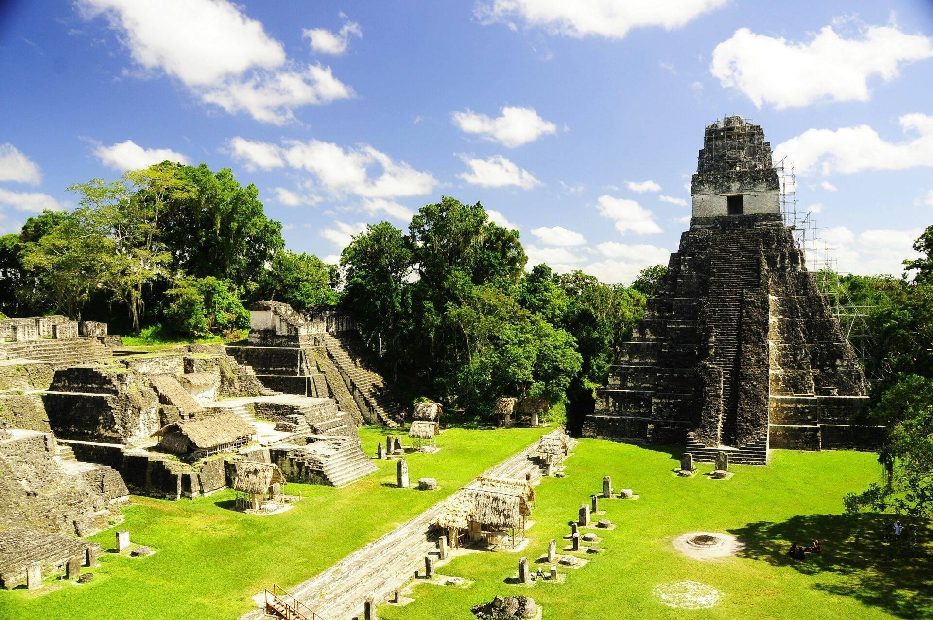 Antike Maya-Ruinen mit zentraler Pyramide, Steinbauten und üppiger tropischer Vegetation unter blauem Himmel