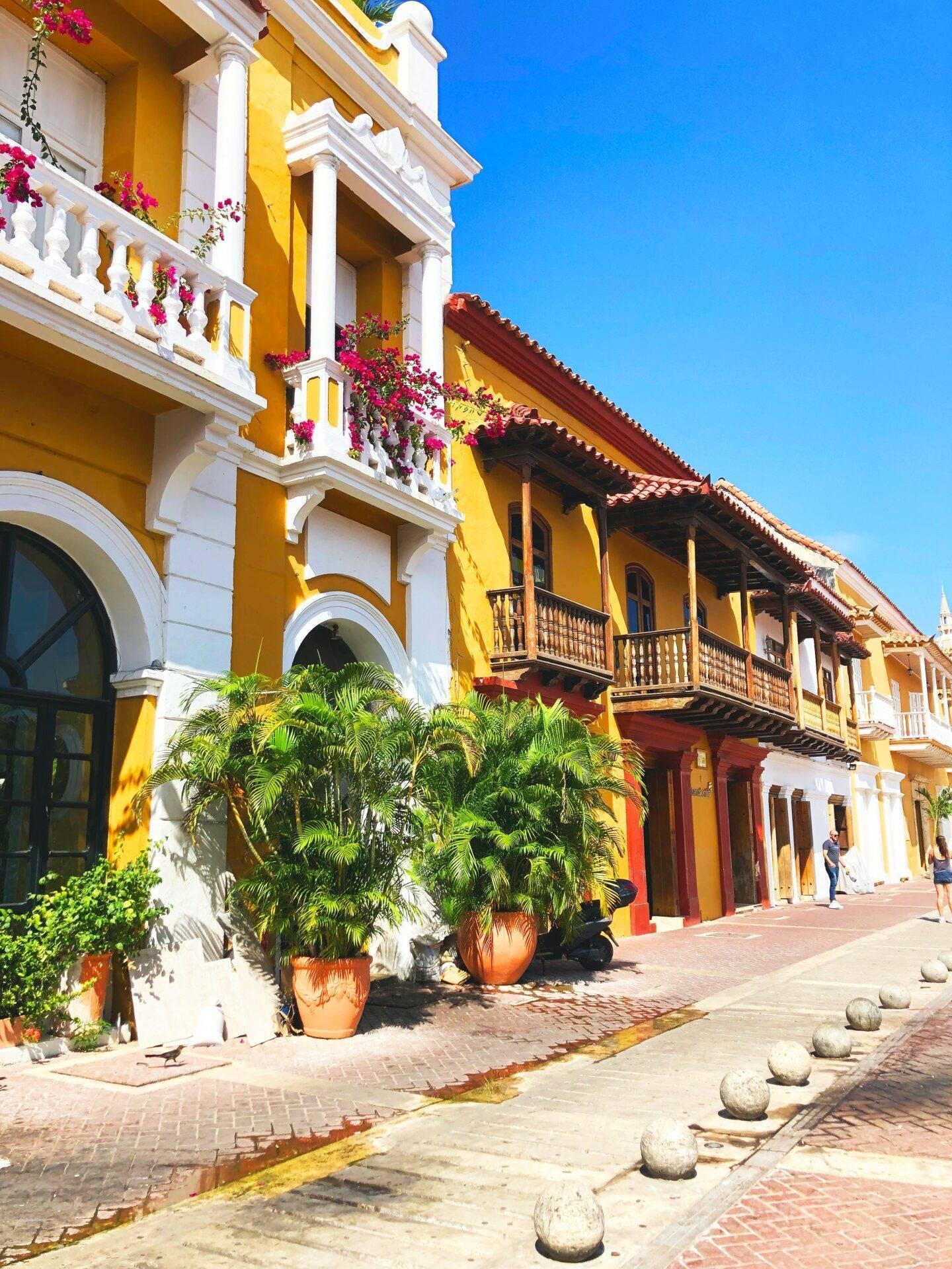 Calle colonial colorida con fachadas amarillas y rojas, balcones de madera, flores magenta y arquitectura hispánica tradicional