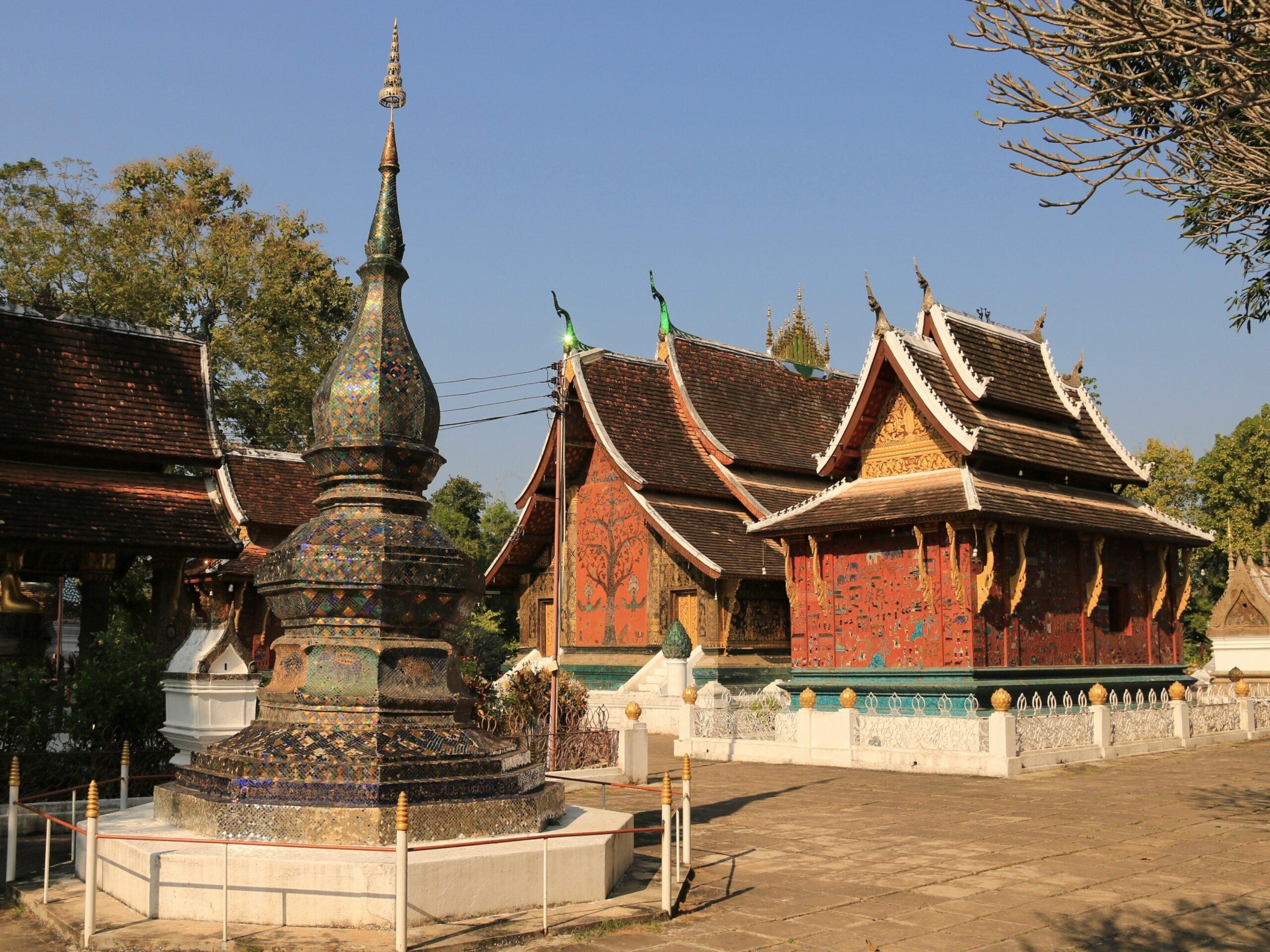 Ein traditioneller buddhistischer Tempel mit geschwungenen goldenen Dächern, verziertem Stupa und roten Gebäuden, umgeben von Bäumen