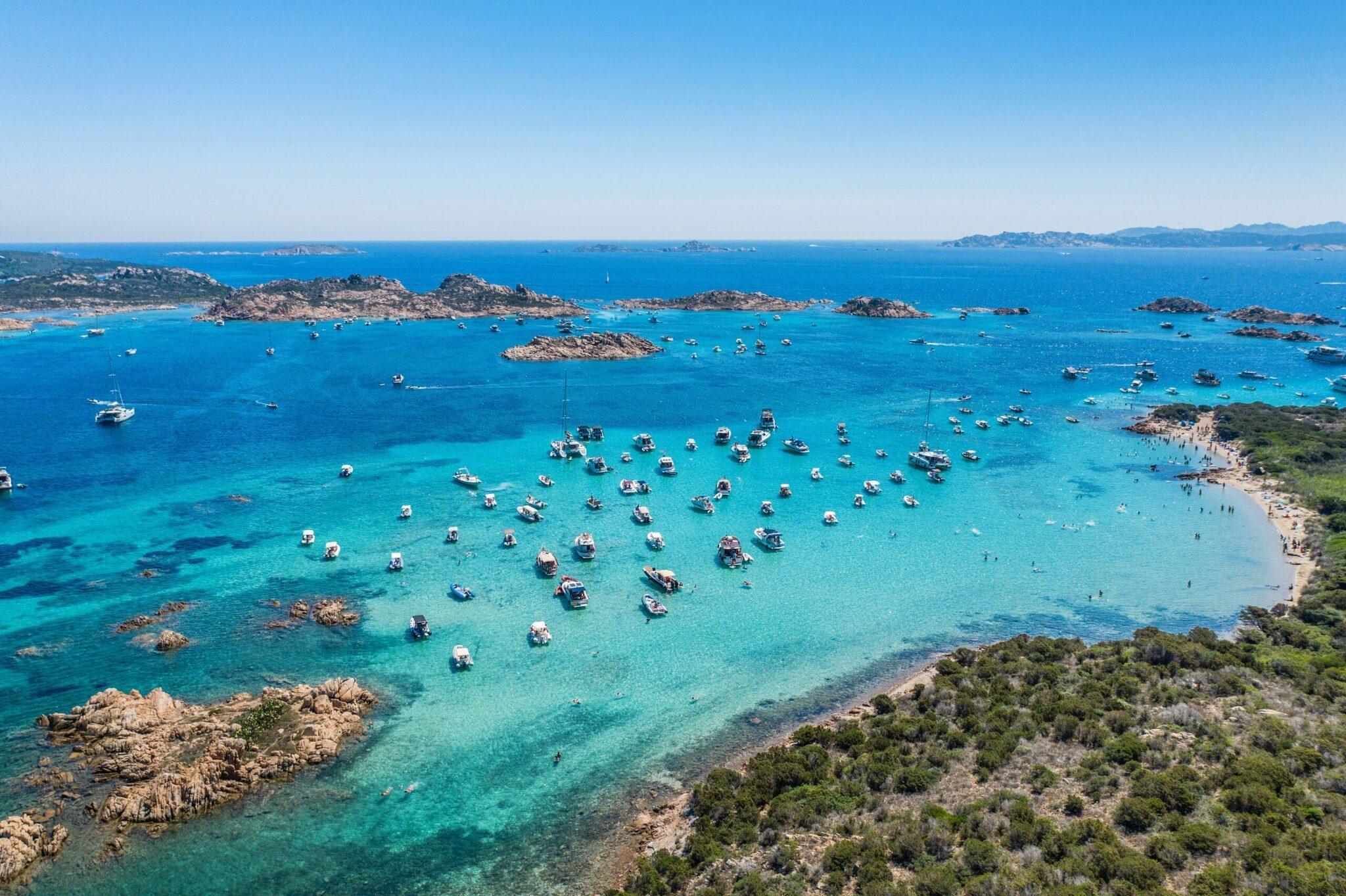 Vista aérea de una bahía turquesa salpicada de embarcaciones fondeadas, rodeada de islotes rocosos y costas mediterráneas de vegetación verde