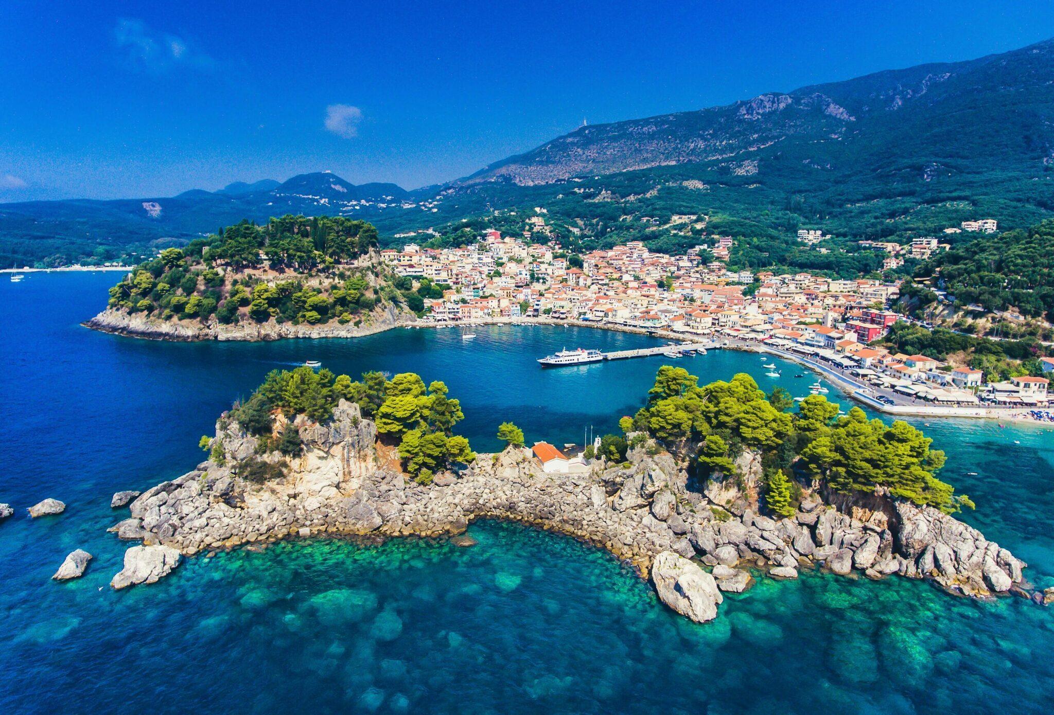 Vista aerea di una baia mediterranea con un isolotto roccioso, una cittadina costiera con tetti arancioni e acque turchesi cristalline