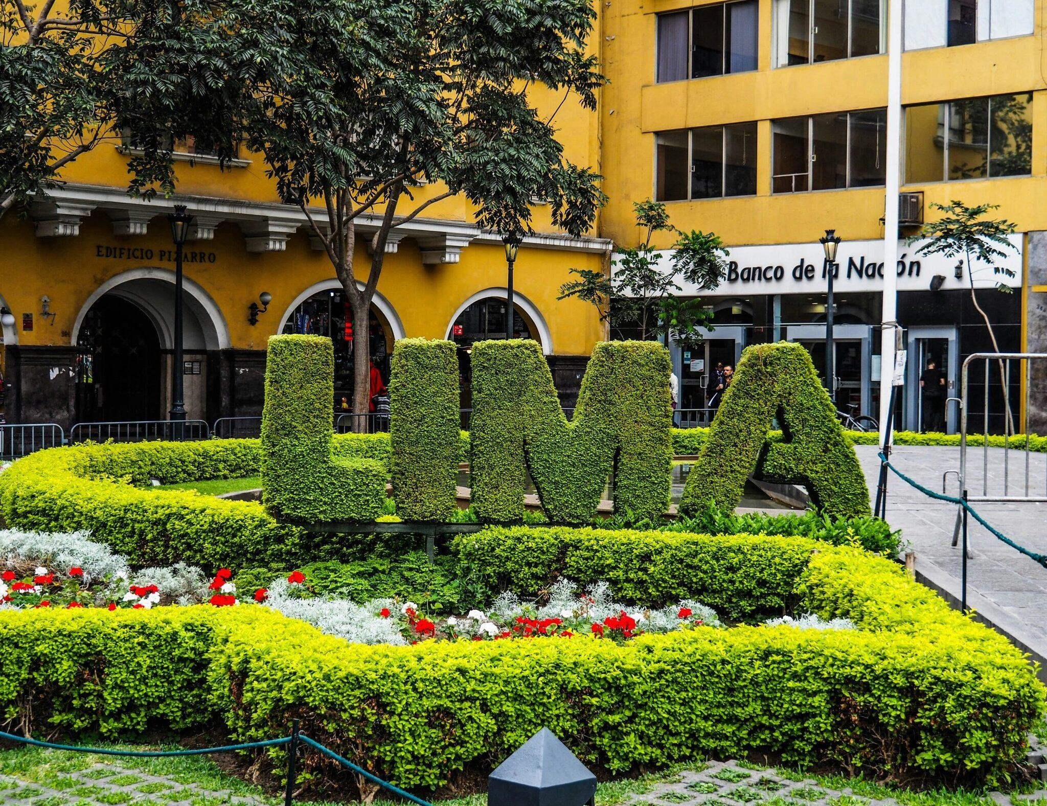 A public square with topiary letter sculptures and flower beds in front of yellow colonial buildings