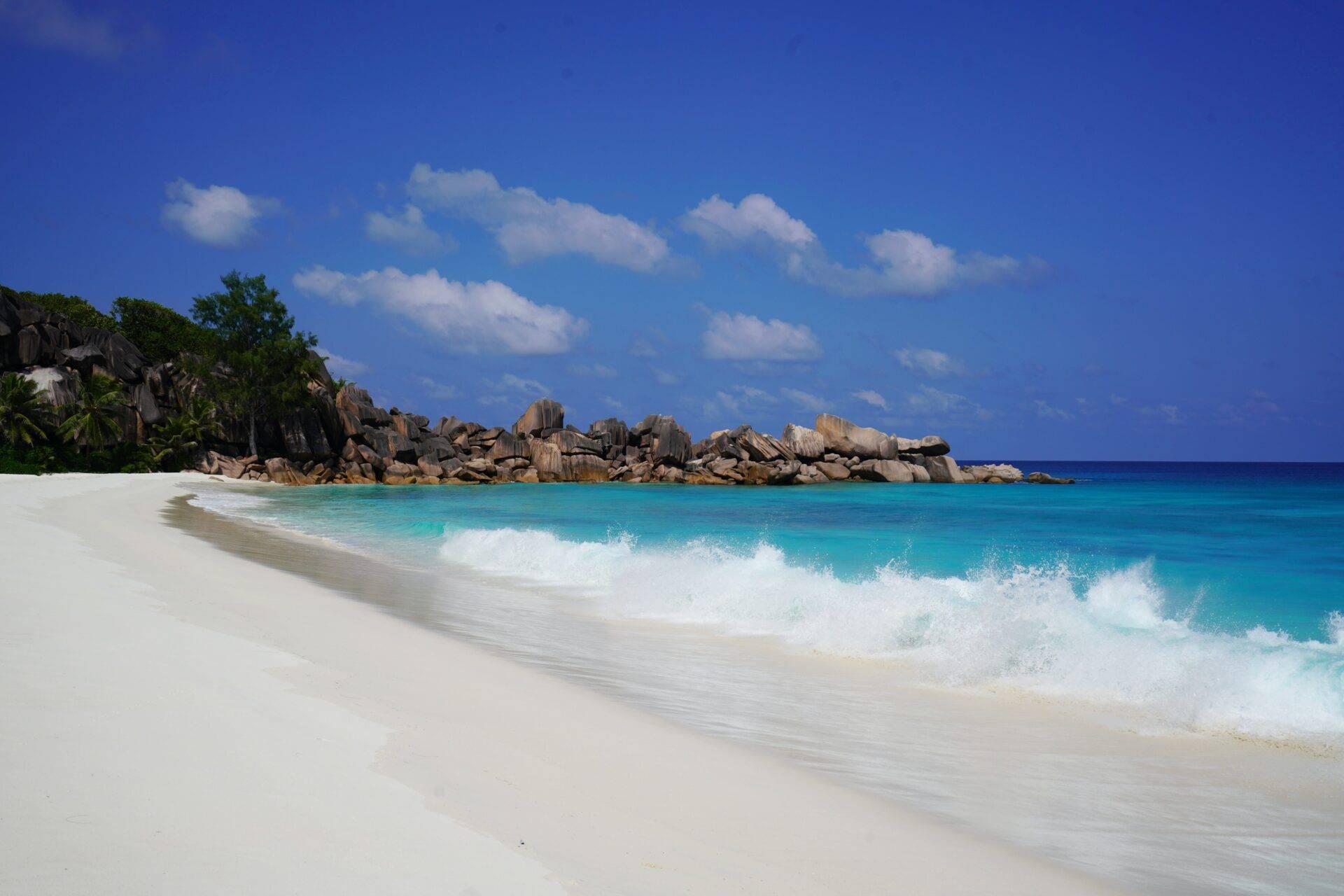 Weißer Sandstrand mit türkisfarbenen Wellen und granitischen Felsformationen unter einem strahlend blauen Himmel