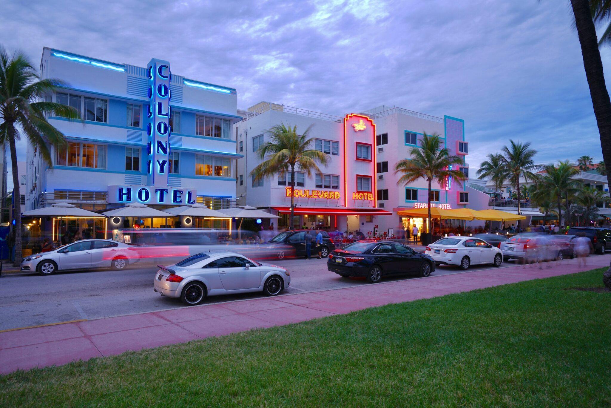 Art Deco hotels lit up at dusk with cars parked along a tropical boulevard