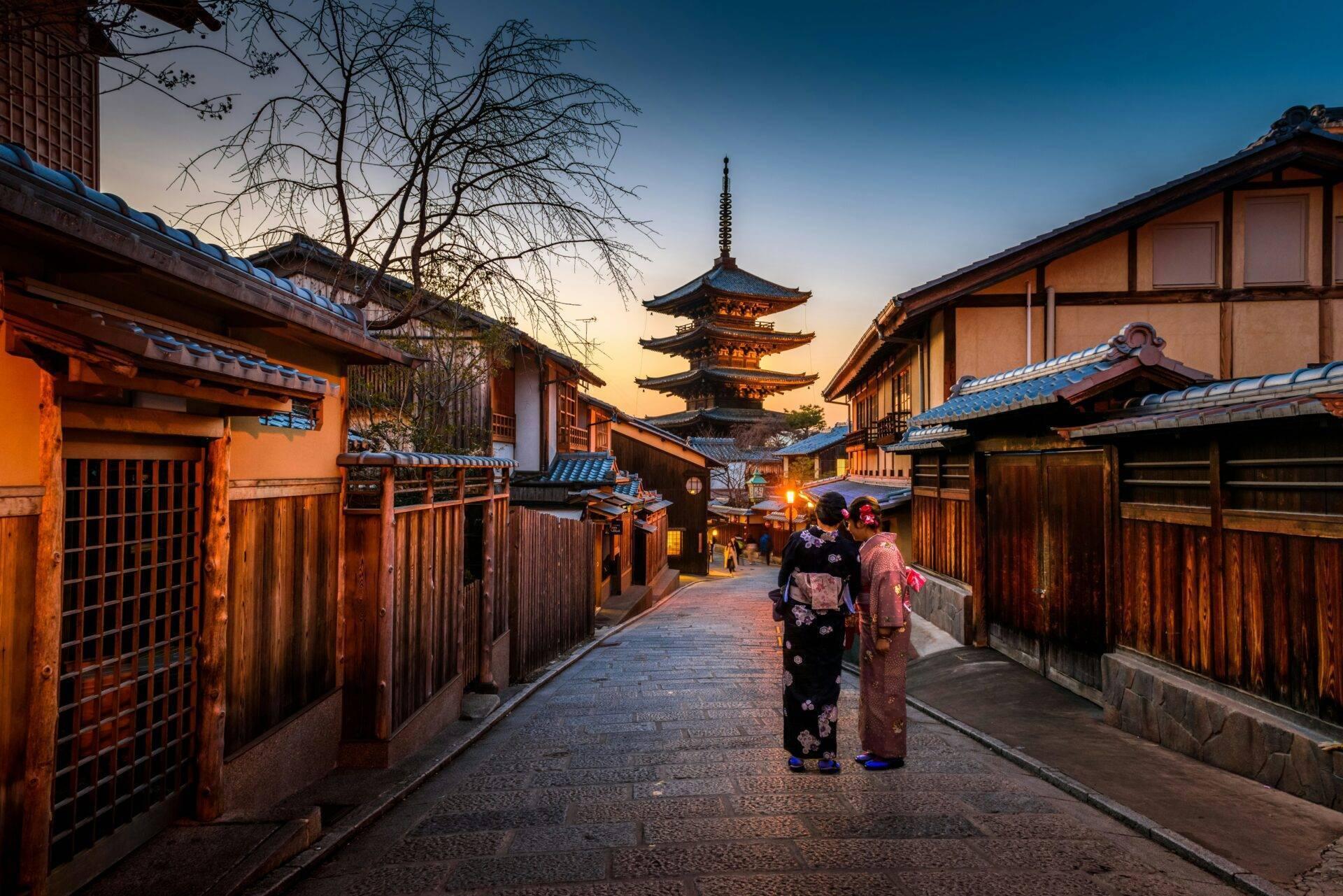 Via storica acciottolata al tramonto con pagoda illuminata, case tradizionali in legno e due donne in kimono