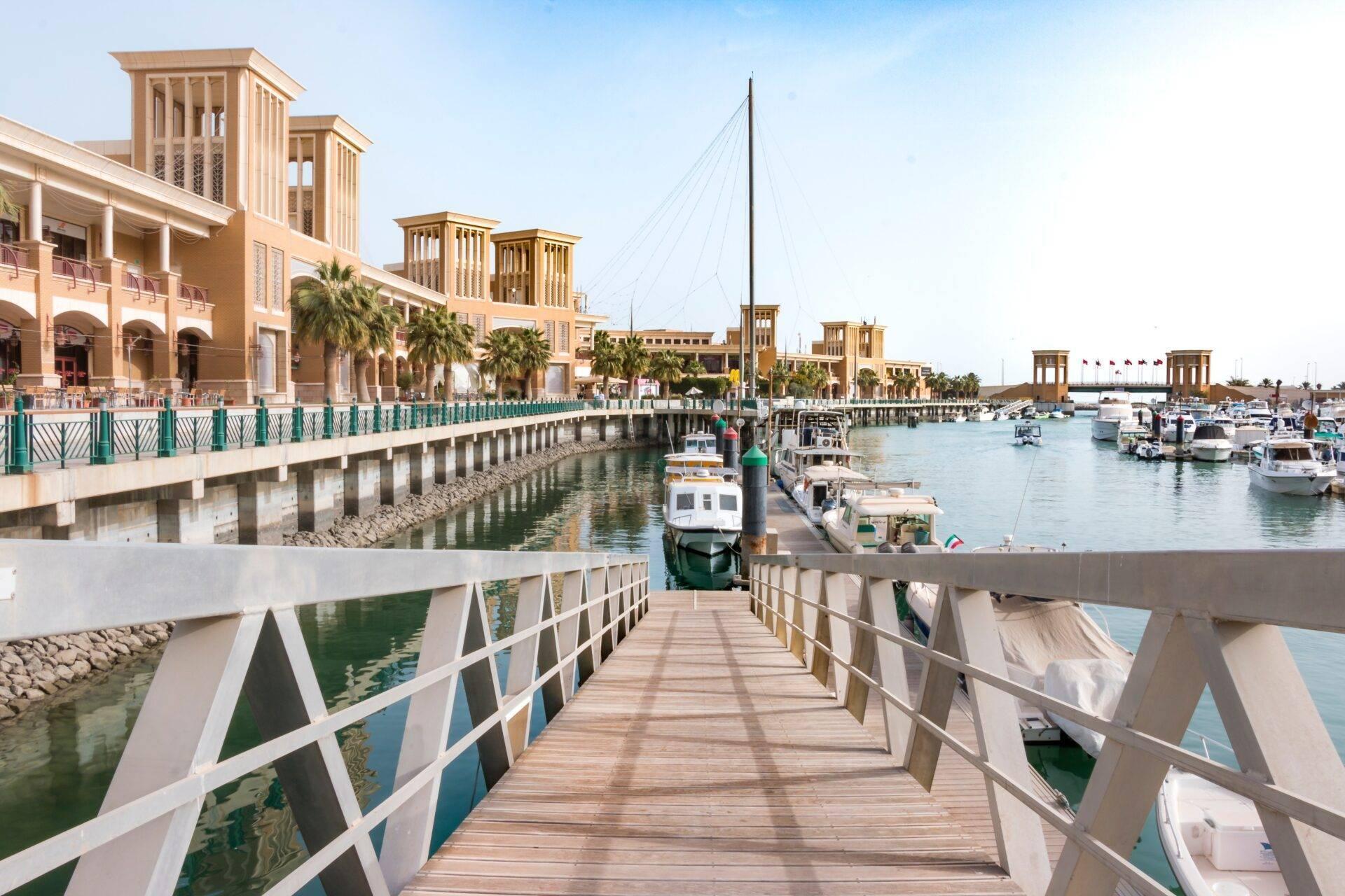 Un embarcadero de madera se adentra en un puerto marina con barcos, edificios tradicionales en beige y palmeras bajo un cielo despejado