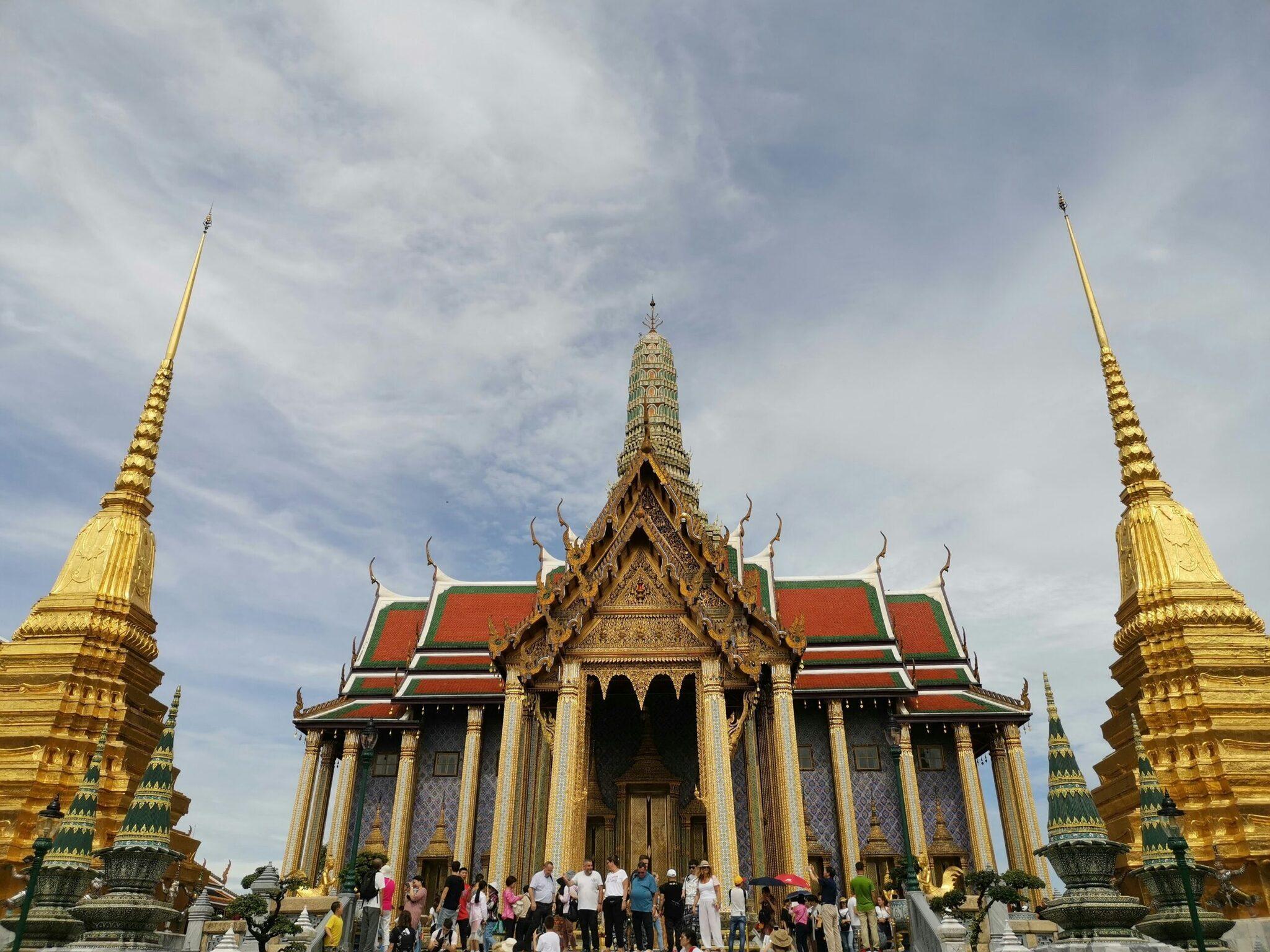 A Thai temple with red roofs and gilded ornaments, with visitors gathered on the main esplanade