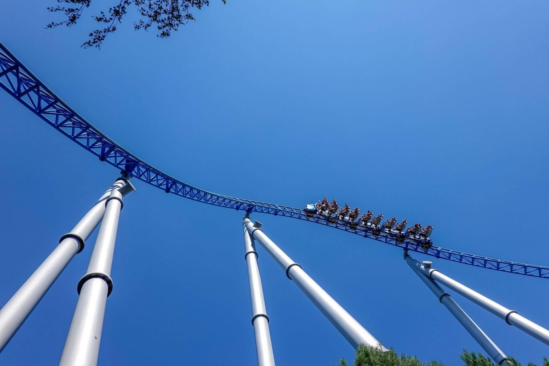 Froschperspektive auf eine Achterbahn mit weißen Trägern und einem Zug voller Fahrgäste unter blauem Himmel