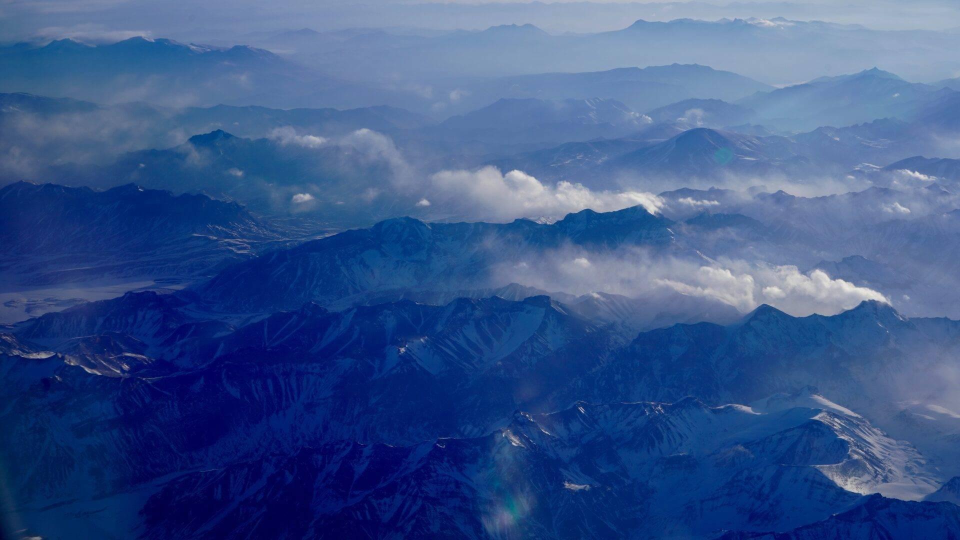 Vista aerea di una grande catena montuosa con cime innevate, vallate profonde e nuvole bianche sparse