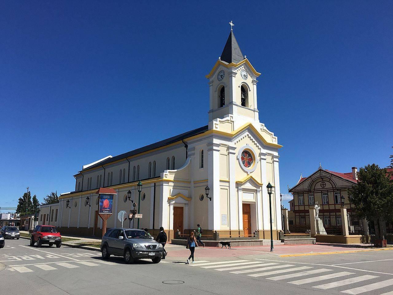 Iglesia blanca con campanario dorado y cruz, plaza pavimentada con peatones y coches, arquitectura colonial sudamericana