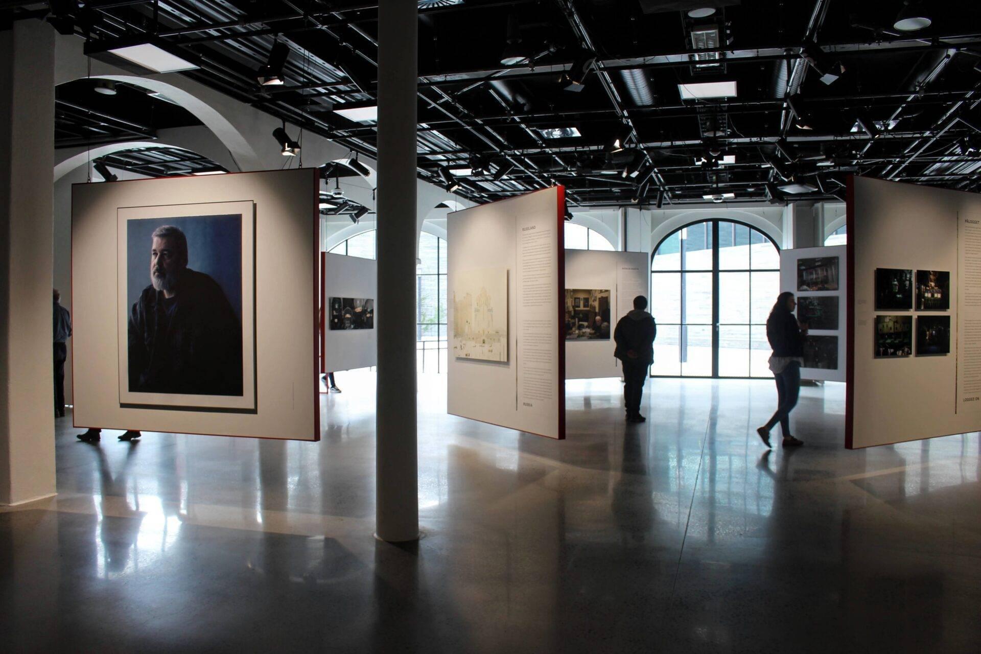 Innenraum einer zeitgenössischen Kunstgalerie mit Besuchern, die Porträts und fotografische Installationen auf weißen Tafeln betrachten