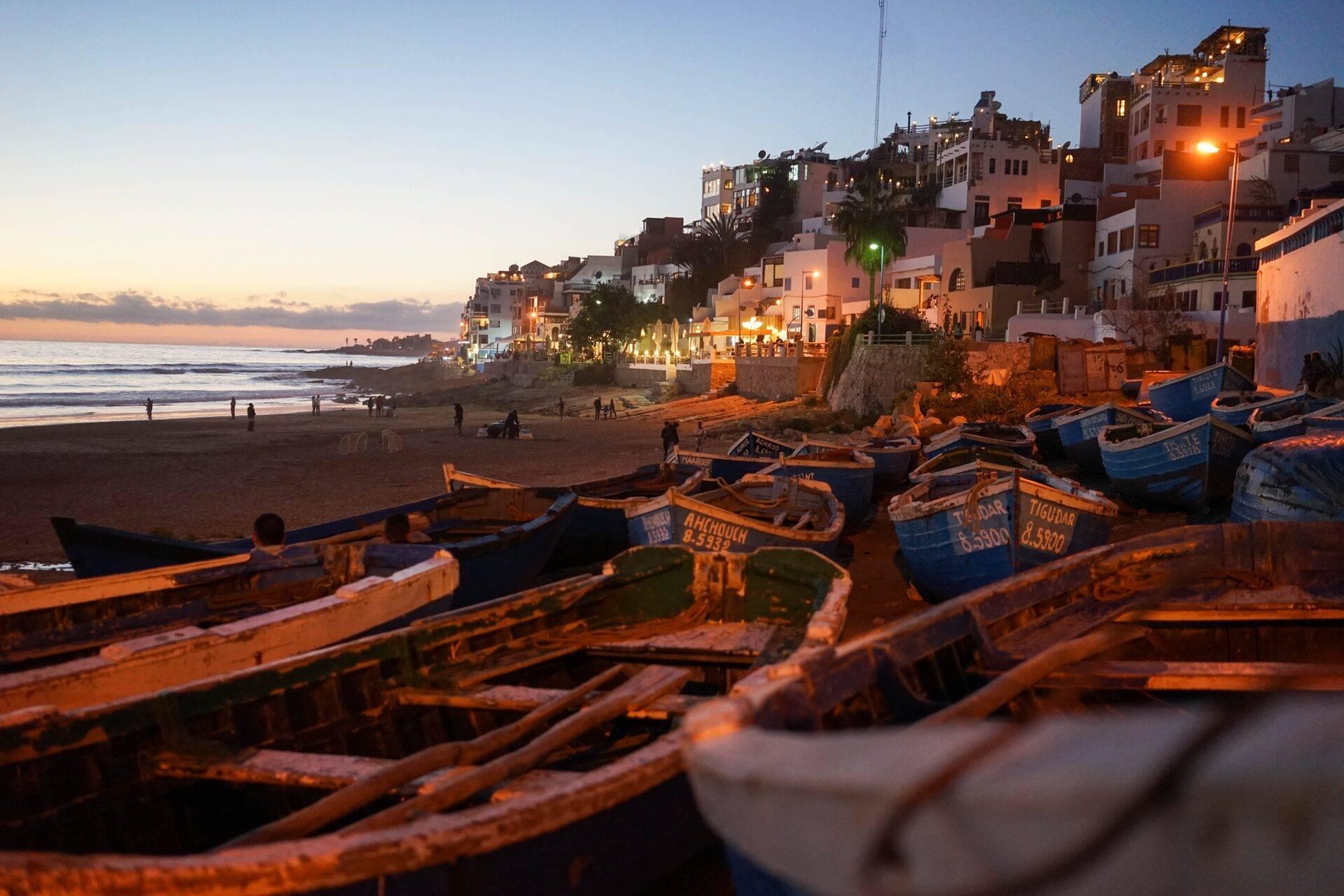 Bunte Fischerboote an einem Strand bei Sonnenuntergang, vor weißen Häusern hoch oben auf der Klippe