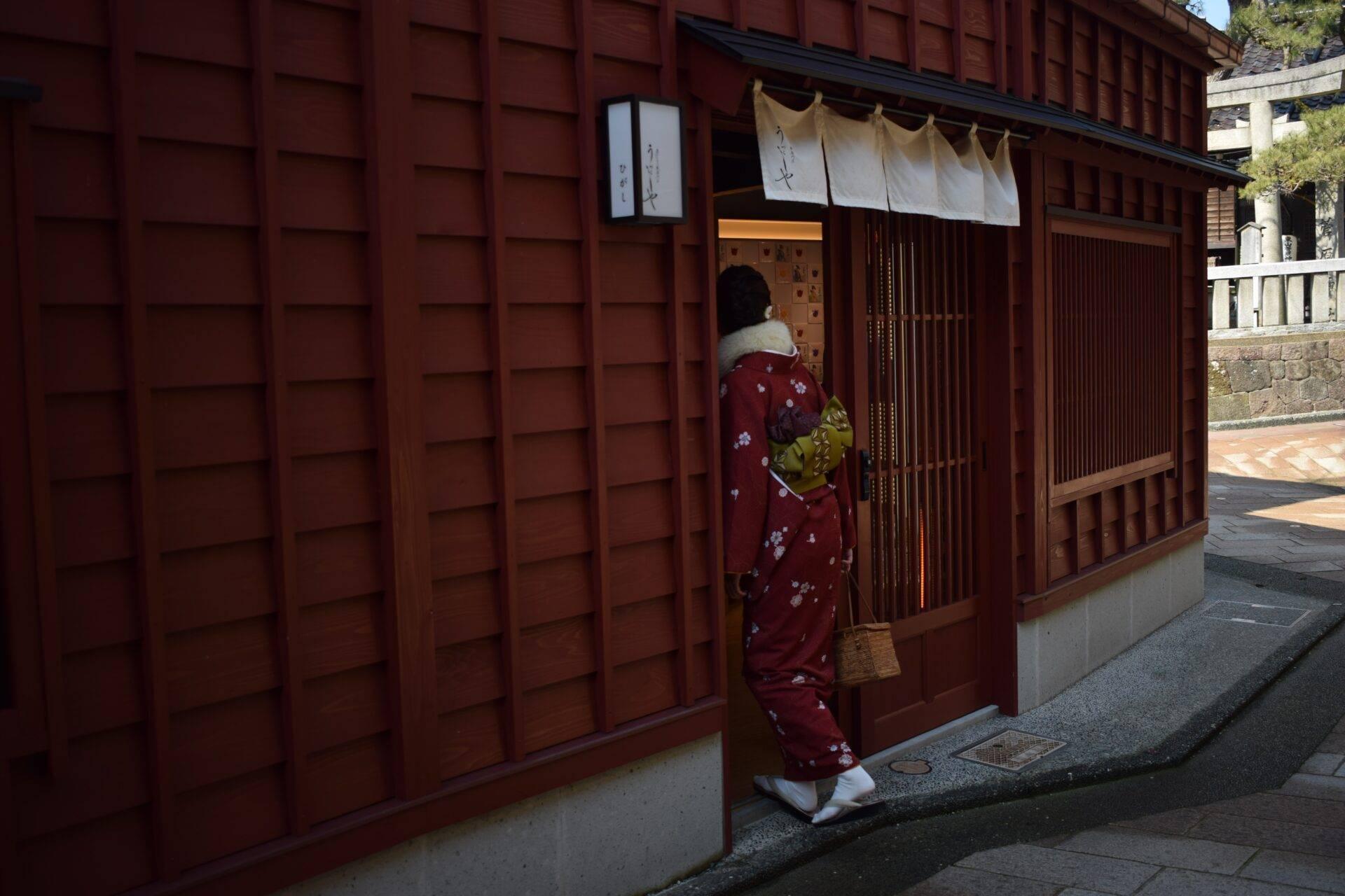 Eine Frau im roten Kimono betritt ein traditionelles japanisches Gebäude mit dunklen Holzwänden und Türen