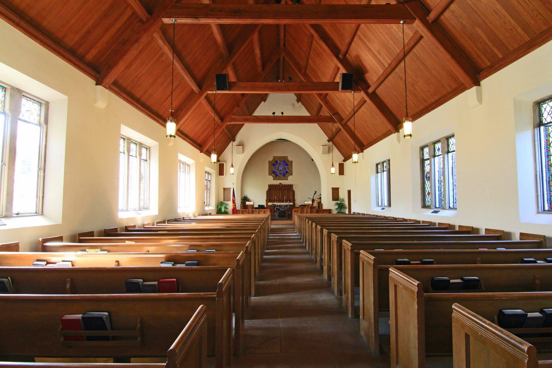 Kircheninnenraum mit Holzbänken, sichtbarem Dachgebälk und farbigen Buntglasfenstern