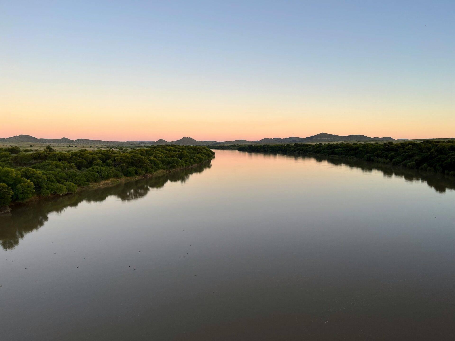 Ein ruhiger Fluss, gesäumt von üppiger Vegetation in der Abenddämmerung, mit felsigen Hügeln im Hintergrund